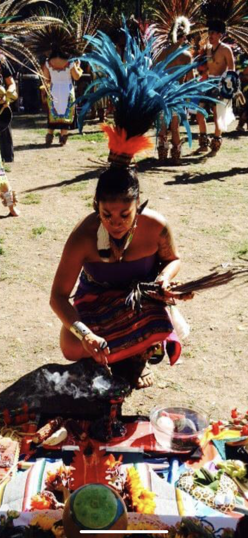A woman in a colorful traditional costume with a large feather headdress participates in a cultural ceremony outdoors, with performers in similar attire and colorful accessories in the background.