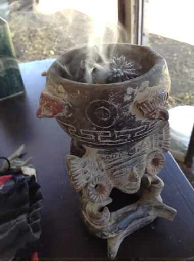 A black chalice filled with smoking incense, placed on a decorated cloth with colorful embroidered patterns, accompanied by a small clay bowl with white stones, a clay pot with painted designs, and a wooden tool with a candle on the side.
