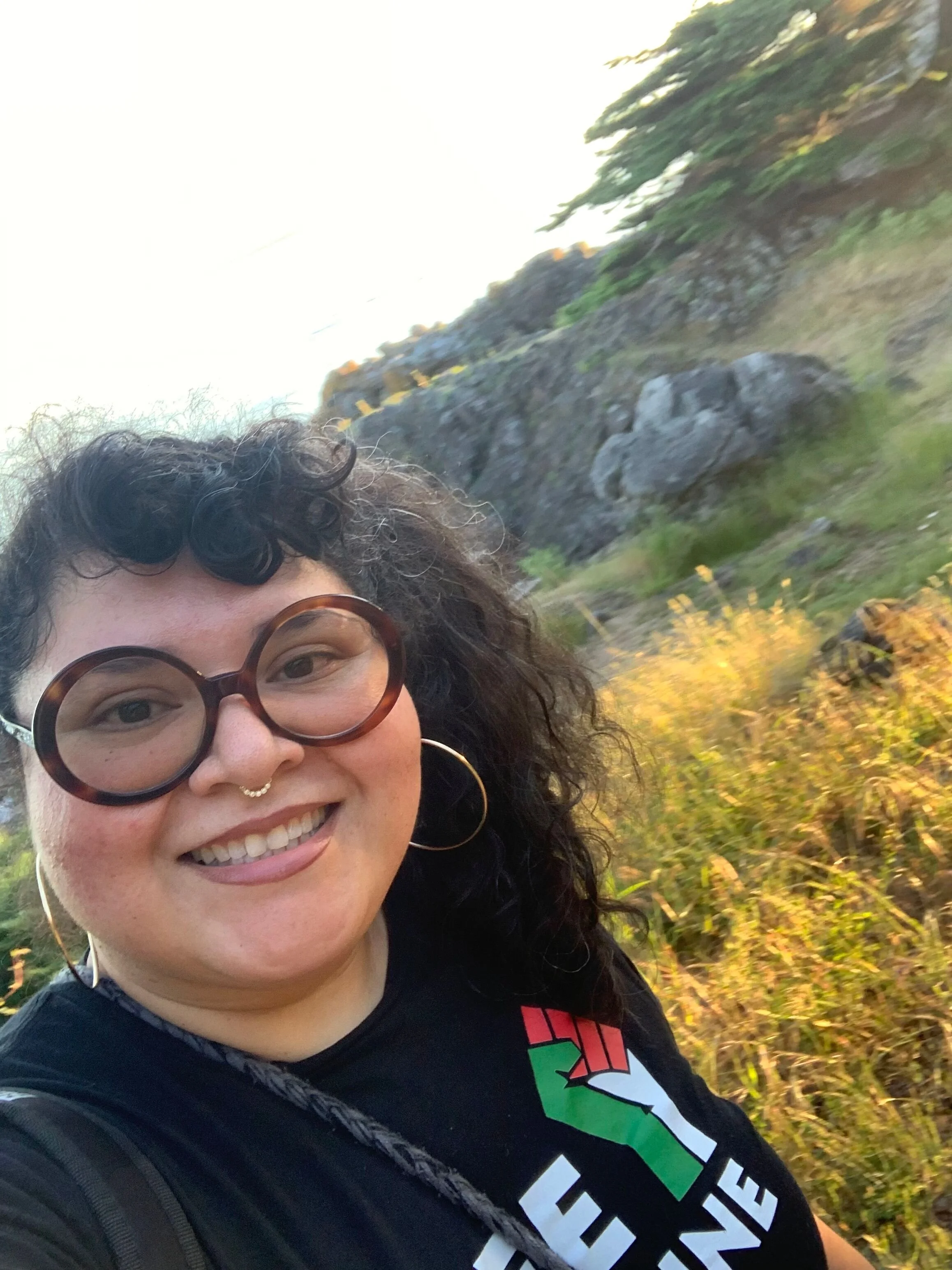 A woman with curly dark hair and large glasses smiling outdoors, with rocks and yellowish foliage in the background during daytime.