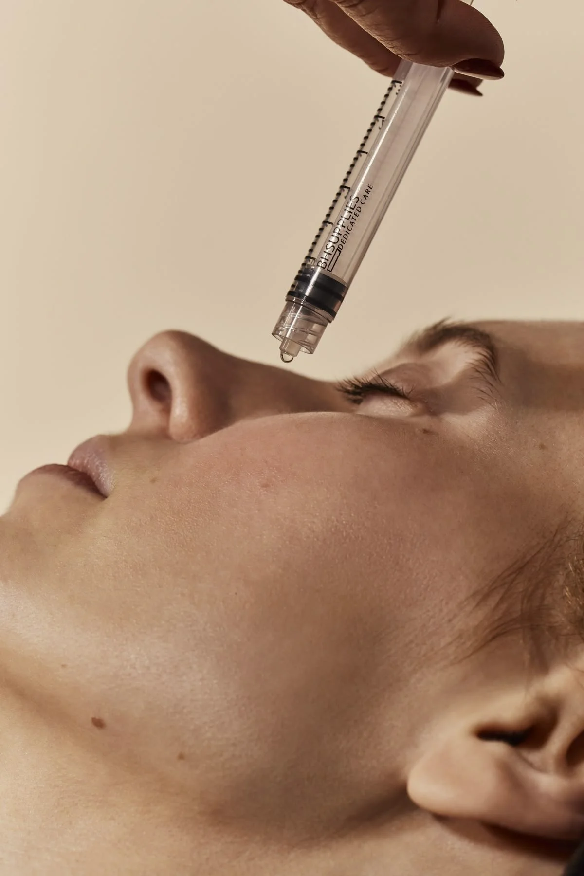 A close-up of a woman receiving a cosmetic treatment, with a syringe held above her face, ready to administer the injection near her eye.