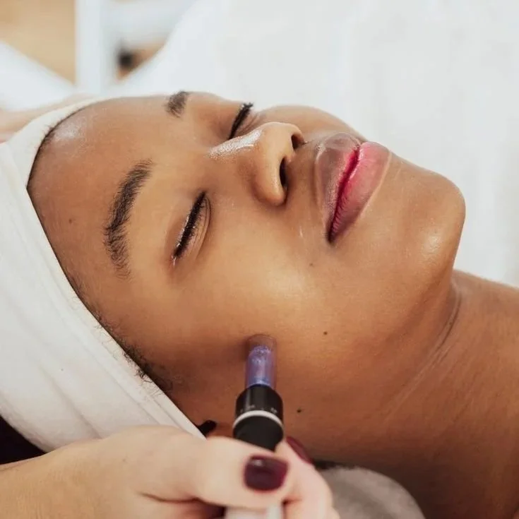 A woman with a headband receiving a facial treatment from a skincare professional using a small device on her cheek.