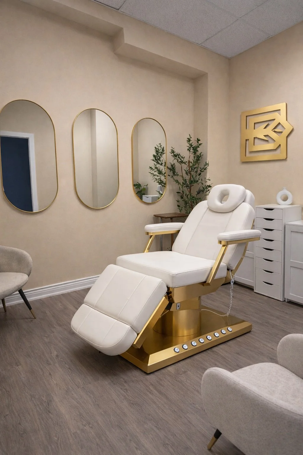 A luxurious medical or spa treatment room with a white reclining chair with gold accents, surrounded by modern decor including three oval mirrors with gold frames on the beige wall, a green plant, a white dresser, and abstract gold wall art.