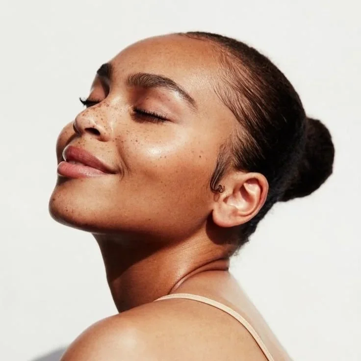Close-up of a young woman with her eyes closed and a content smile, showing freckles on her face and styled hair in a bun, against a plain background.