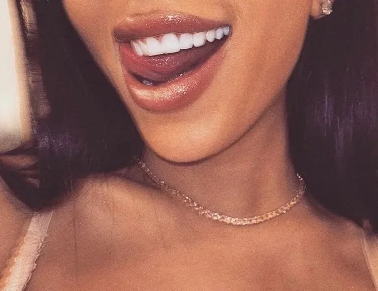 Close-up of a woman smiling and sticking out her tongue, showing white teeth and glossy lips, wearing a delicate necklace.