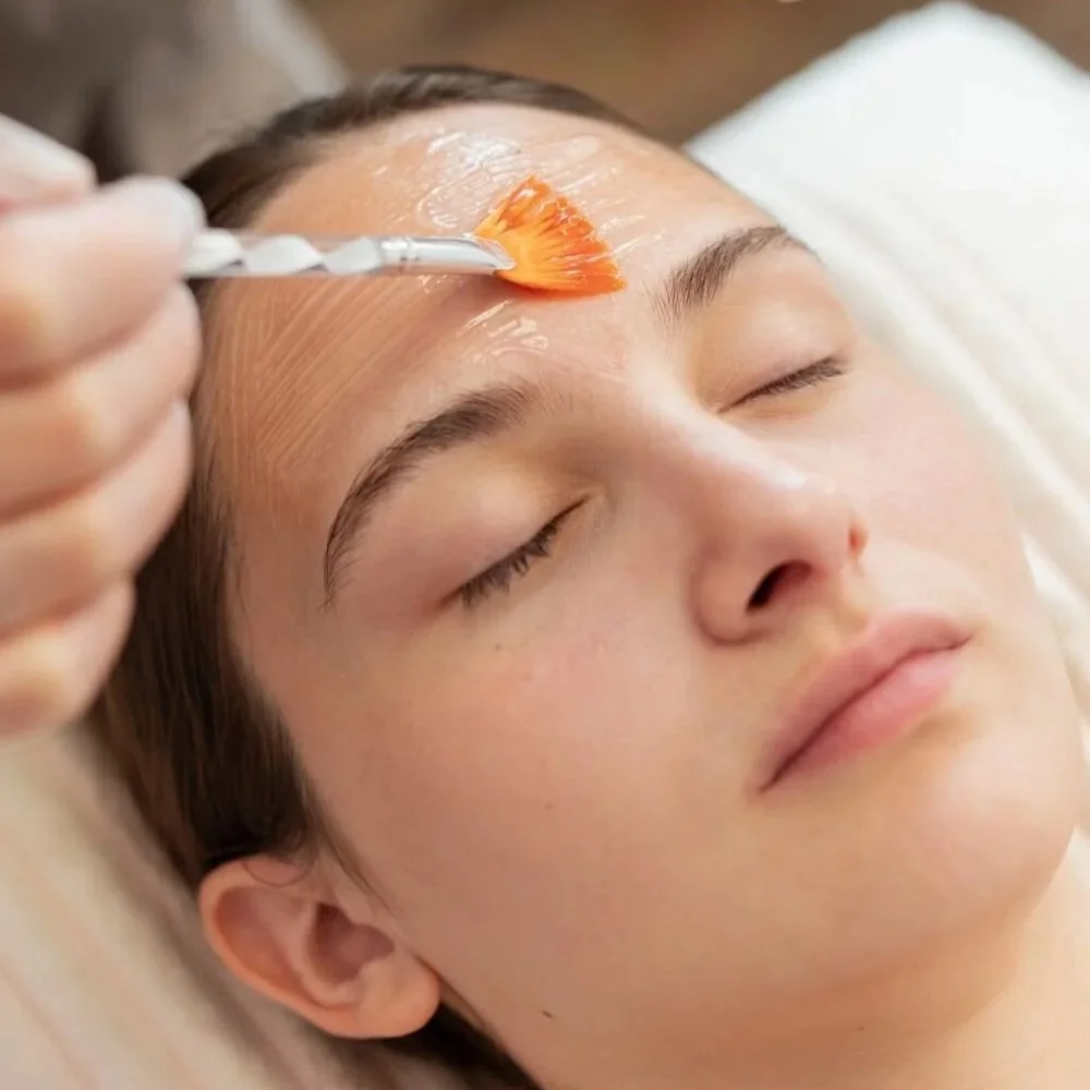 A woman receiving a facial treatment, with a skincare product applied to her forehead using a spatula.