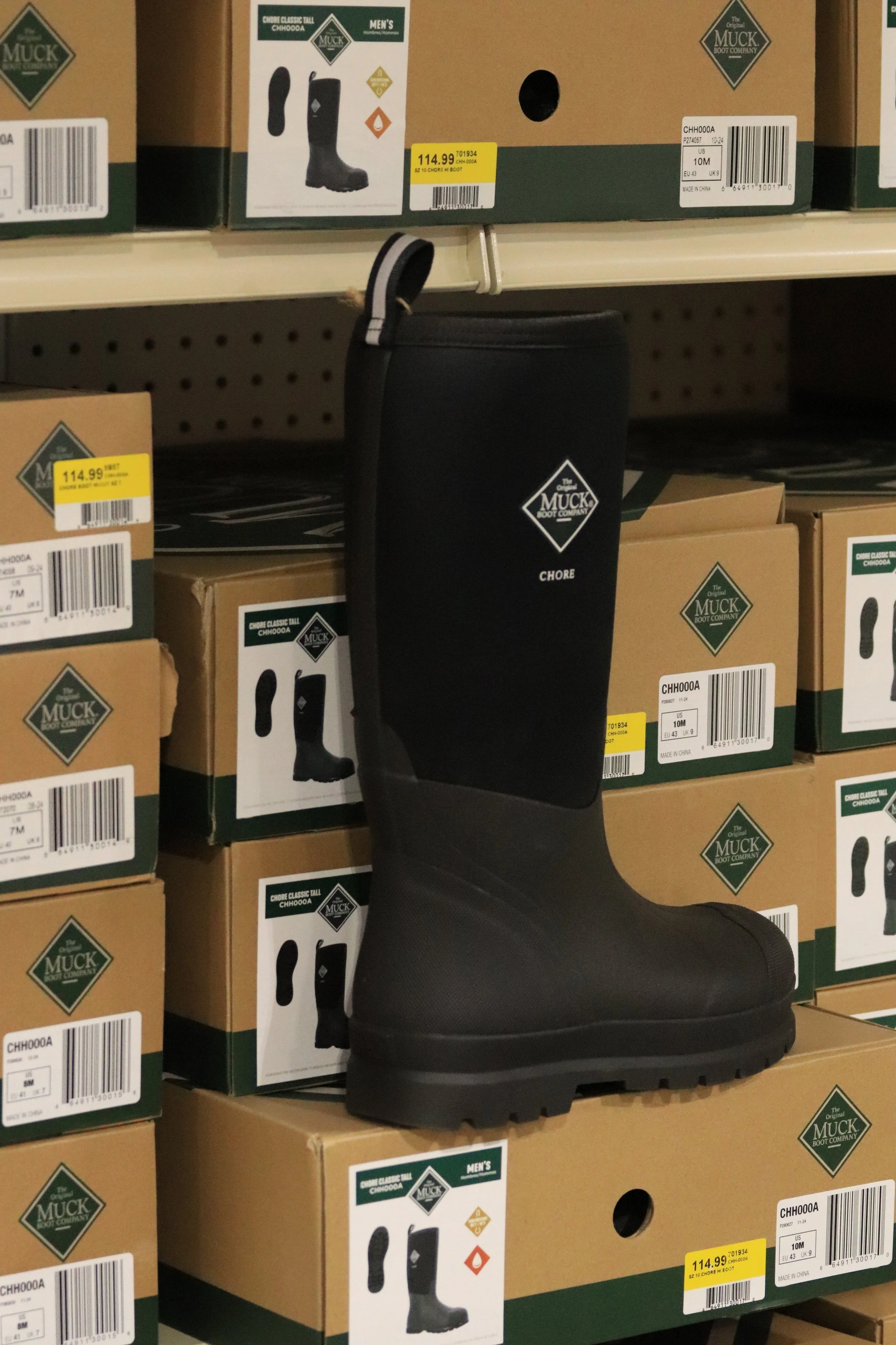 Black Muck Boot Company Chore tall rubber work boot displayed on store shelf, surrounded by similar boxed boots.