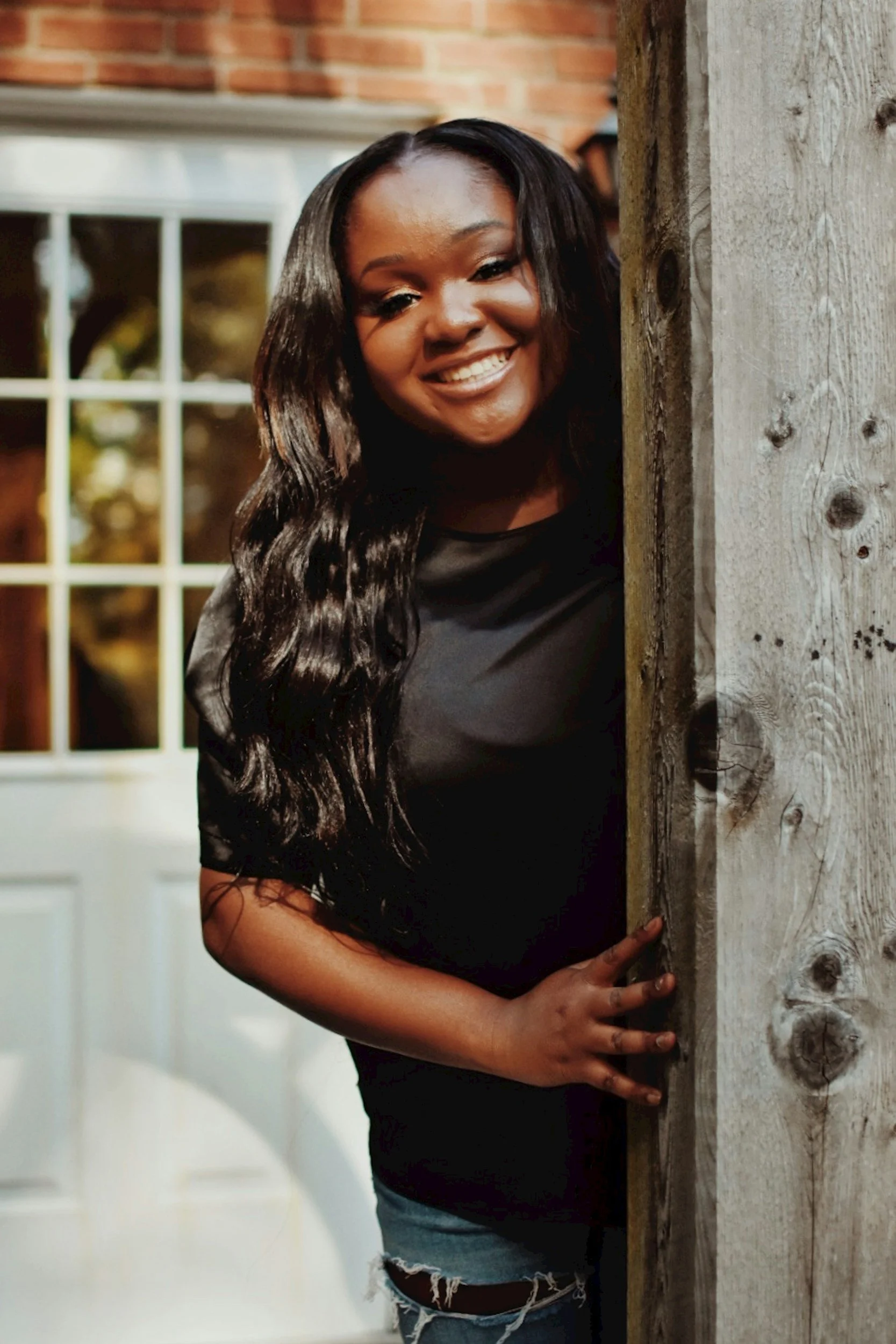 Smiling Black woman with long dark hair standing behind a rustic wooden door outside a brick house