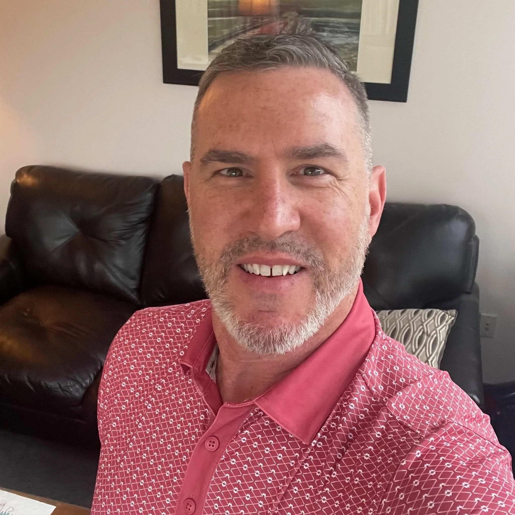 Smiling middle-aged man in a patterned pink polo shirt taking a selfie on a leather couch