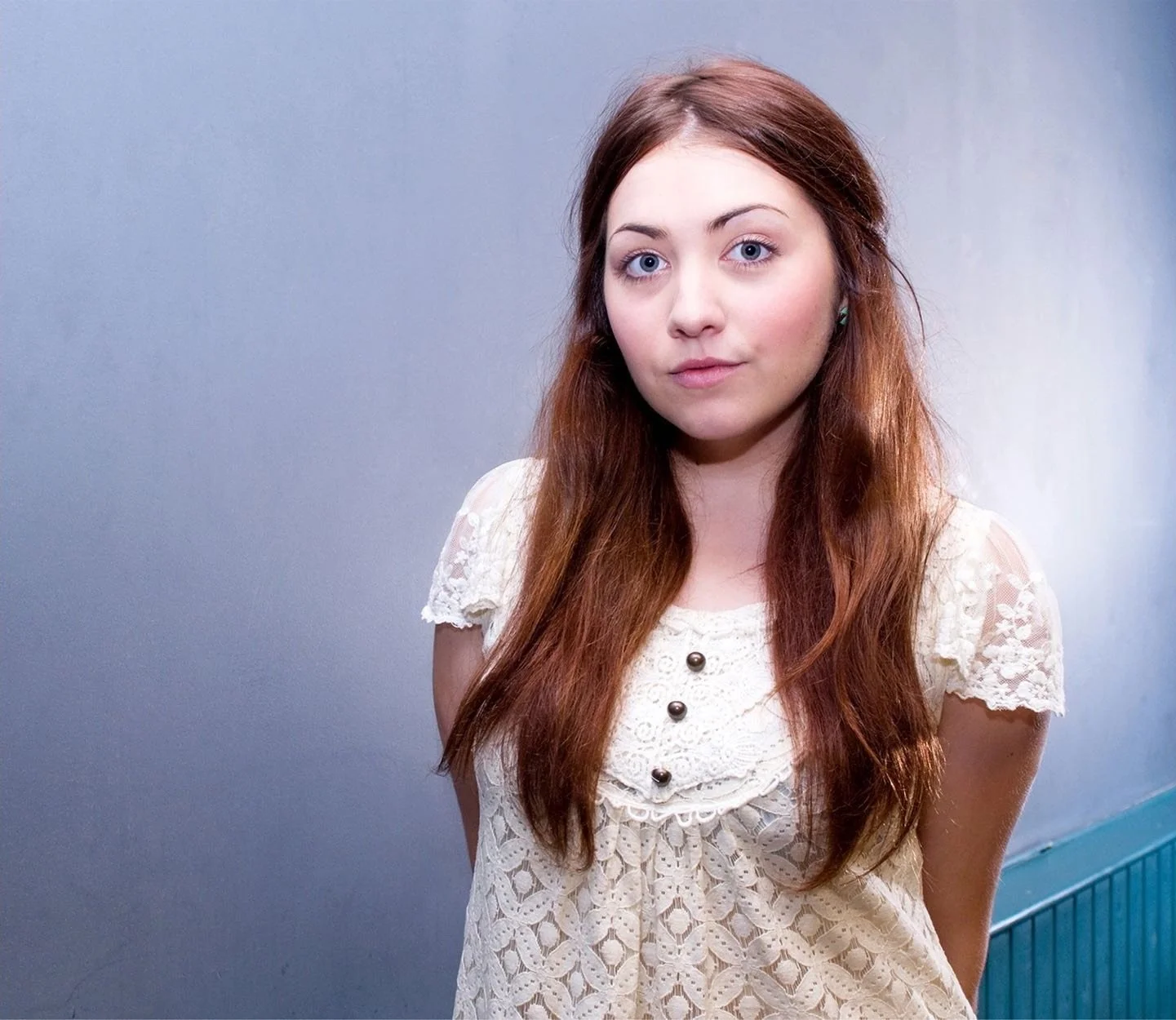 Woman with long brown hair wearing a white lace top standing against a gray background.