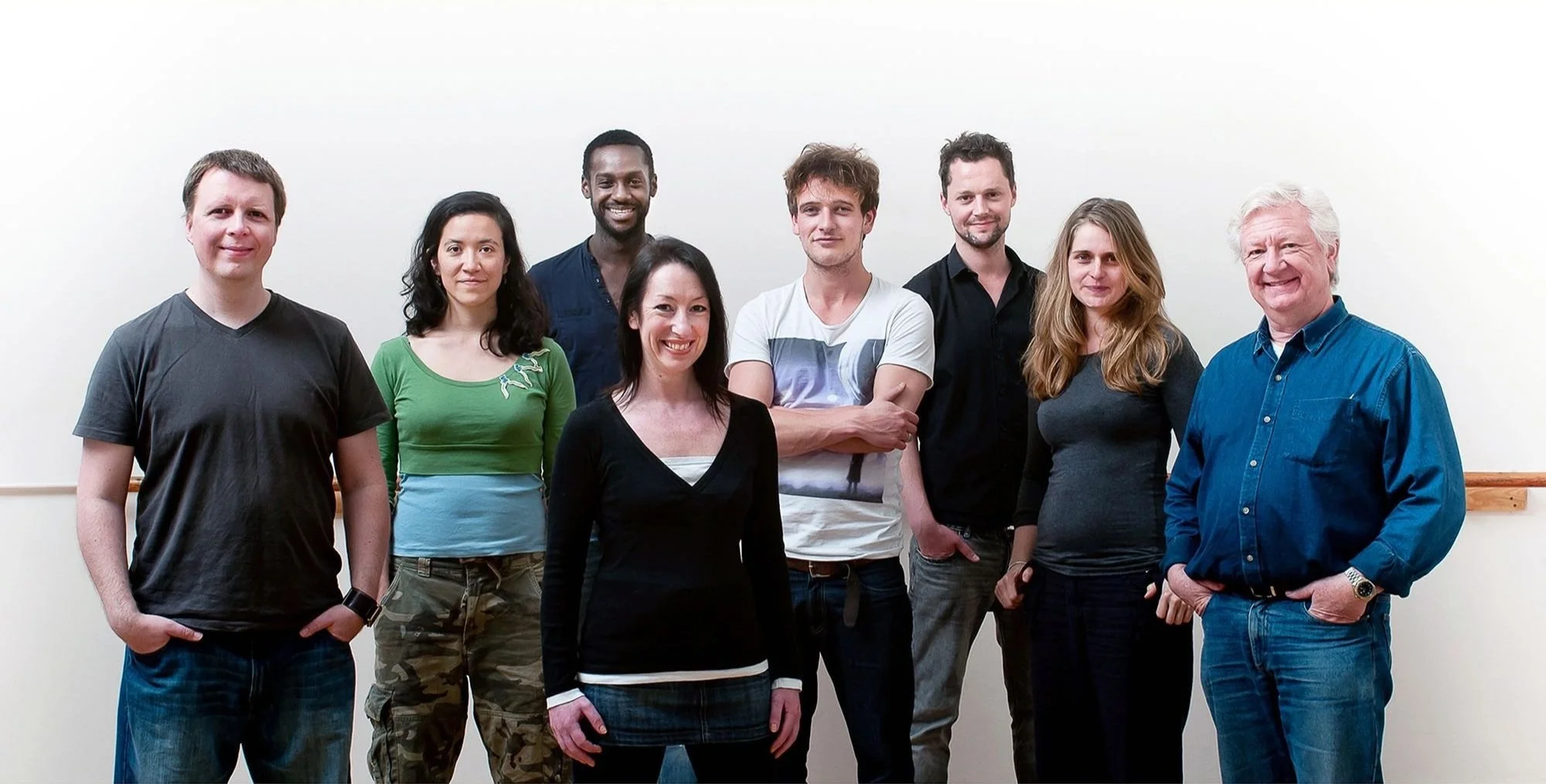Group of eight people smiling, standing side by side against a plain background. They are casually dressed, with a mix of men and women.