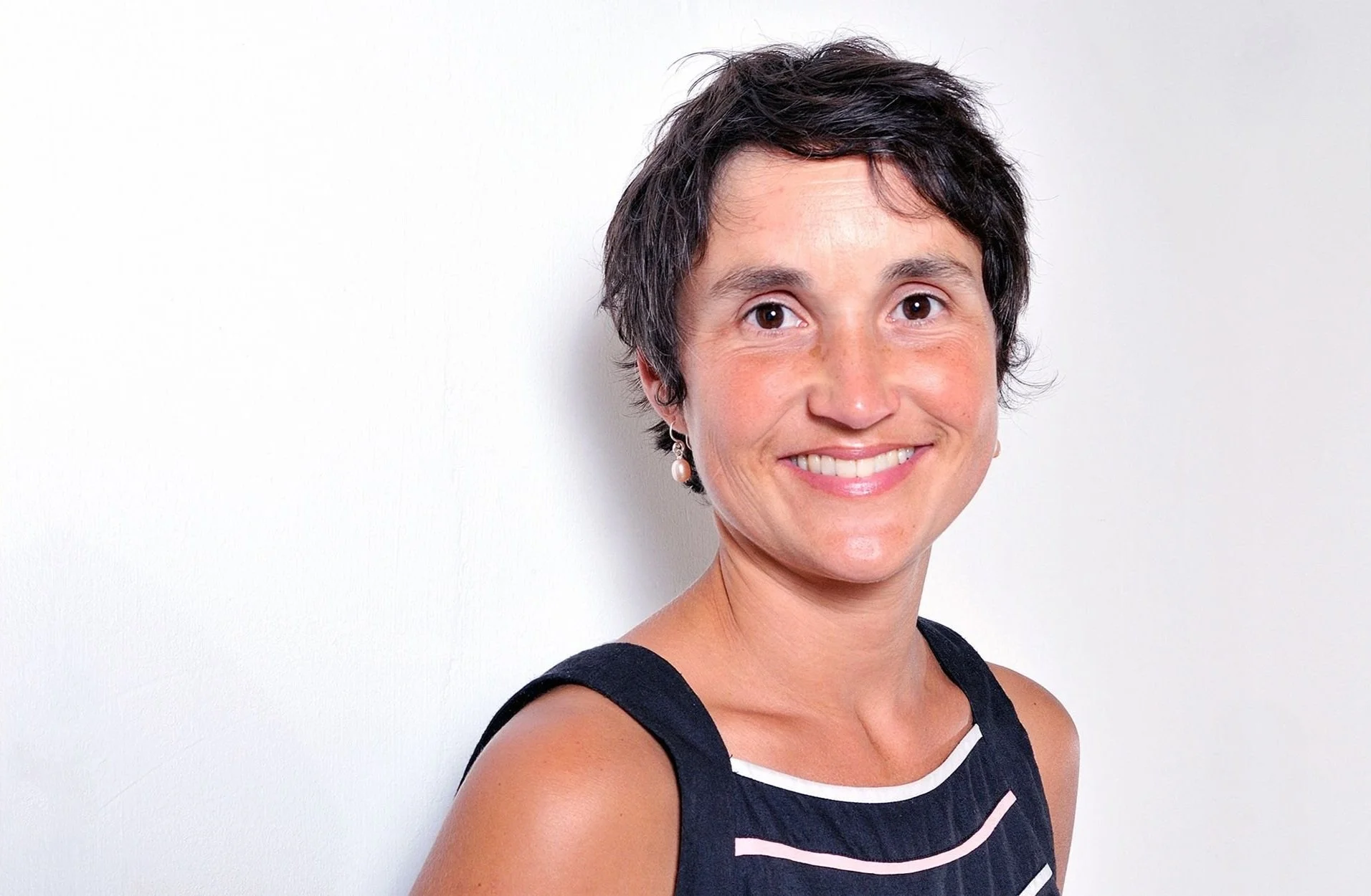 Smiling woman with short dark hair, wearing a sleeveless top, standing against a plain white background.