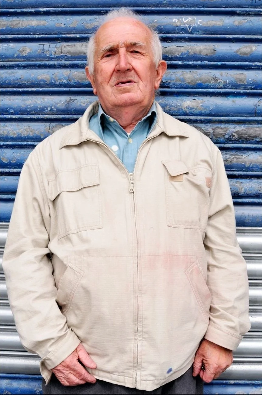 Elderly man in beige jacket standing against a blue metal shutter.