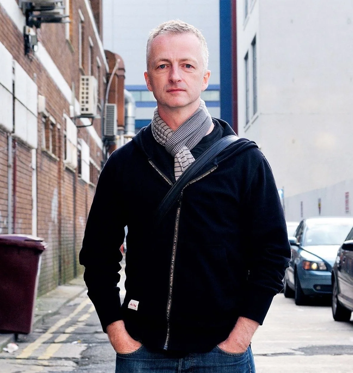 Man standing in an urban alley, wearing a black hoodie and a gray scarf, with hands in pockets and a car in the background.