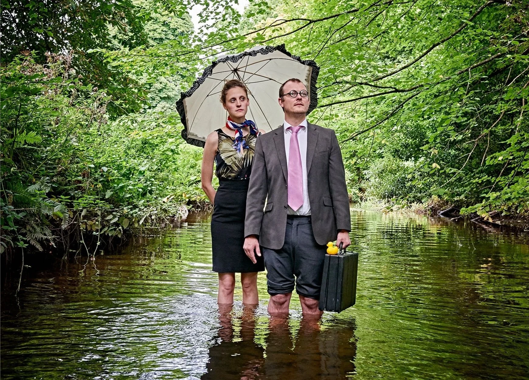 A man and woman standing in a shallow stream; the man is in a suit holding a briefcase and rubber duck, woman holds a parasol, surrounded by lush green foliage.