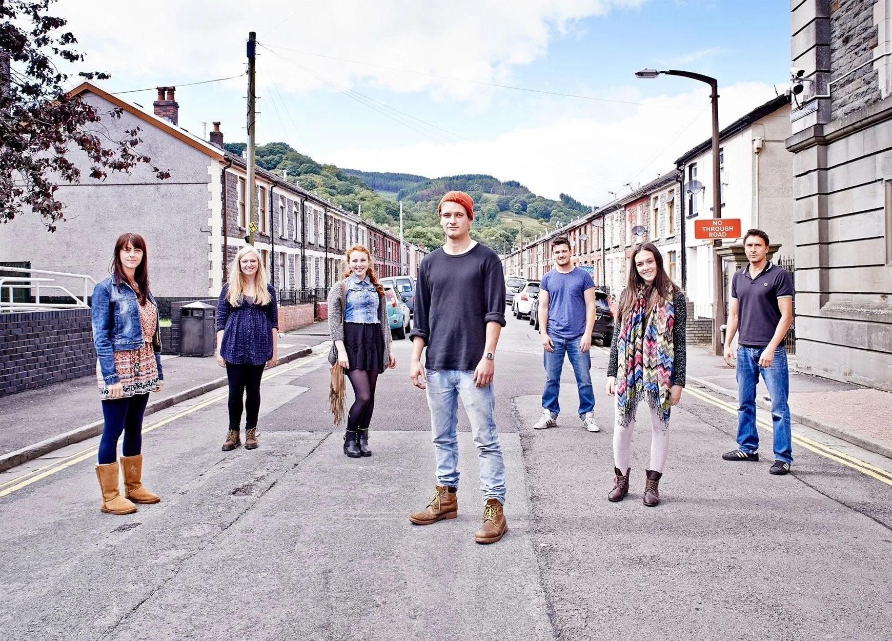 Group of people standing on a residential street with terraced houses in the background, against a backdrop of hills and sky.