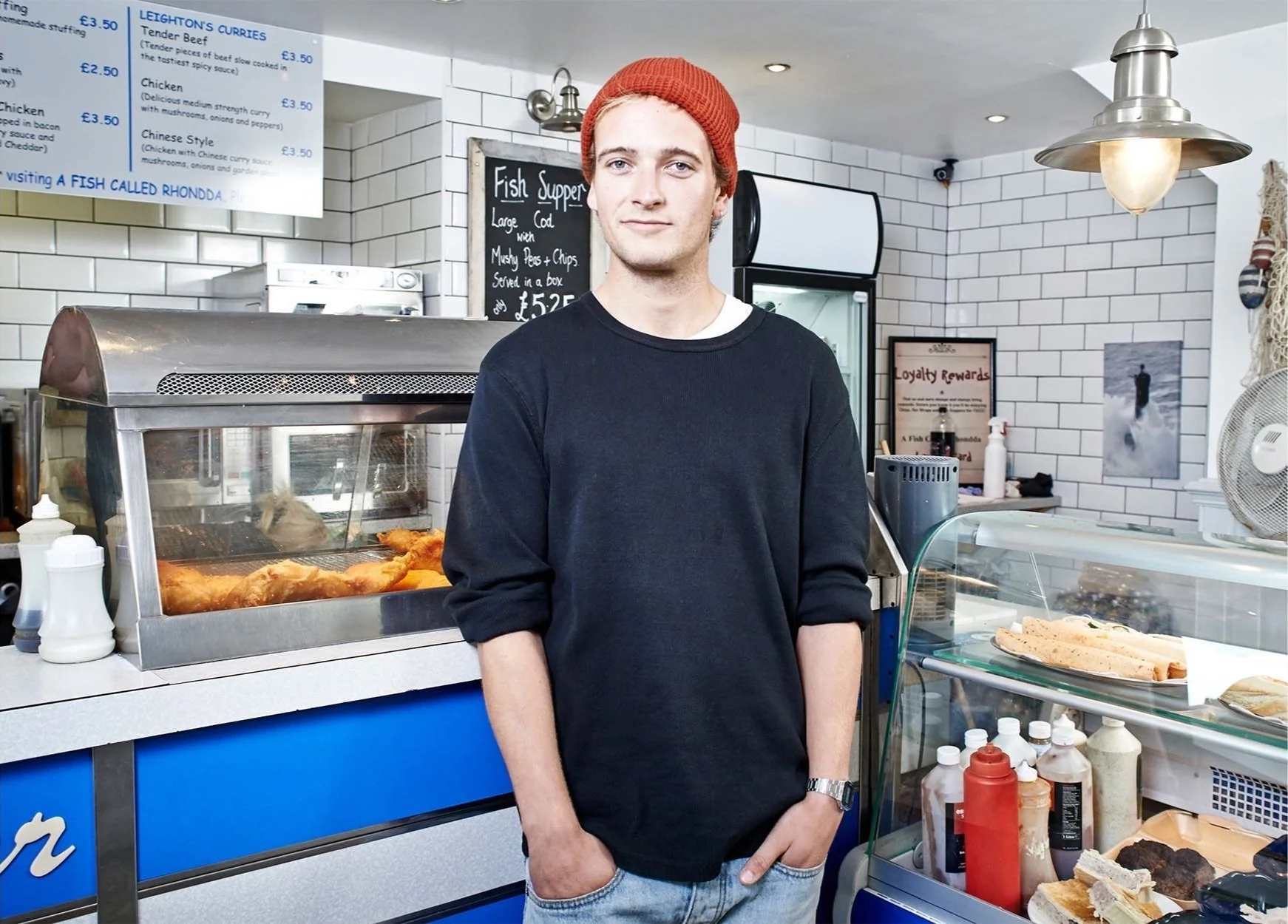 Man wearing a red beanie and black sweater standing inside a fish and chips shop with a blue counter and menu signs.