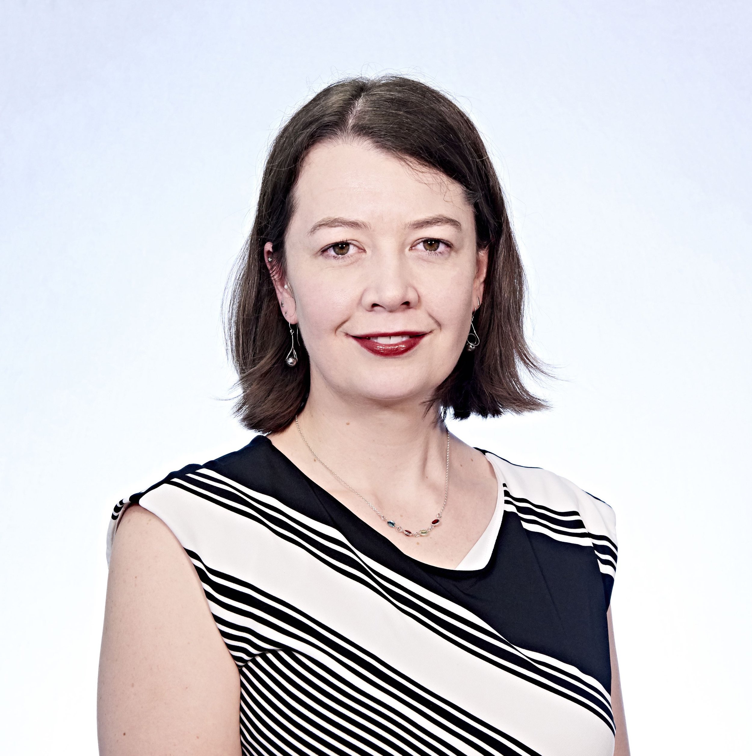 Portrait of a woman with shoulder-length brown hair, wearing a black and white striped dress and earrings, against a light background.