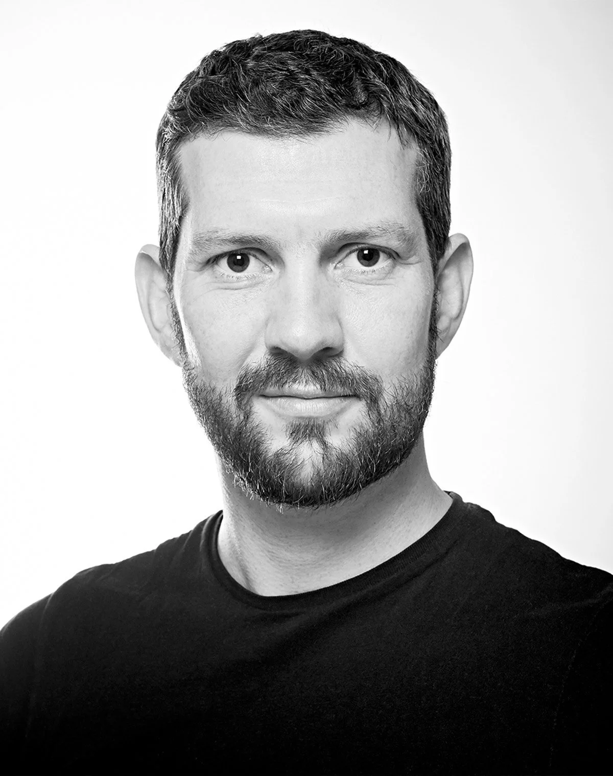 Black and white portrait of a man with short hair and a beard, wearing a dark shirt, looking at the camera.