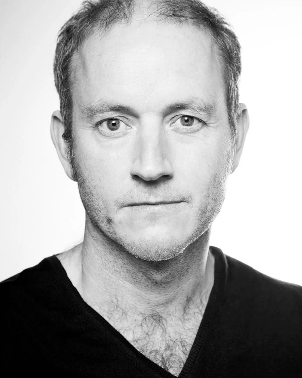 Black and white portrait of a man looking at the camera, with a neutral expression, wearing a V-neck shirt.