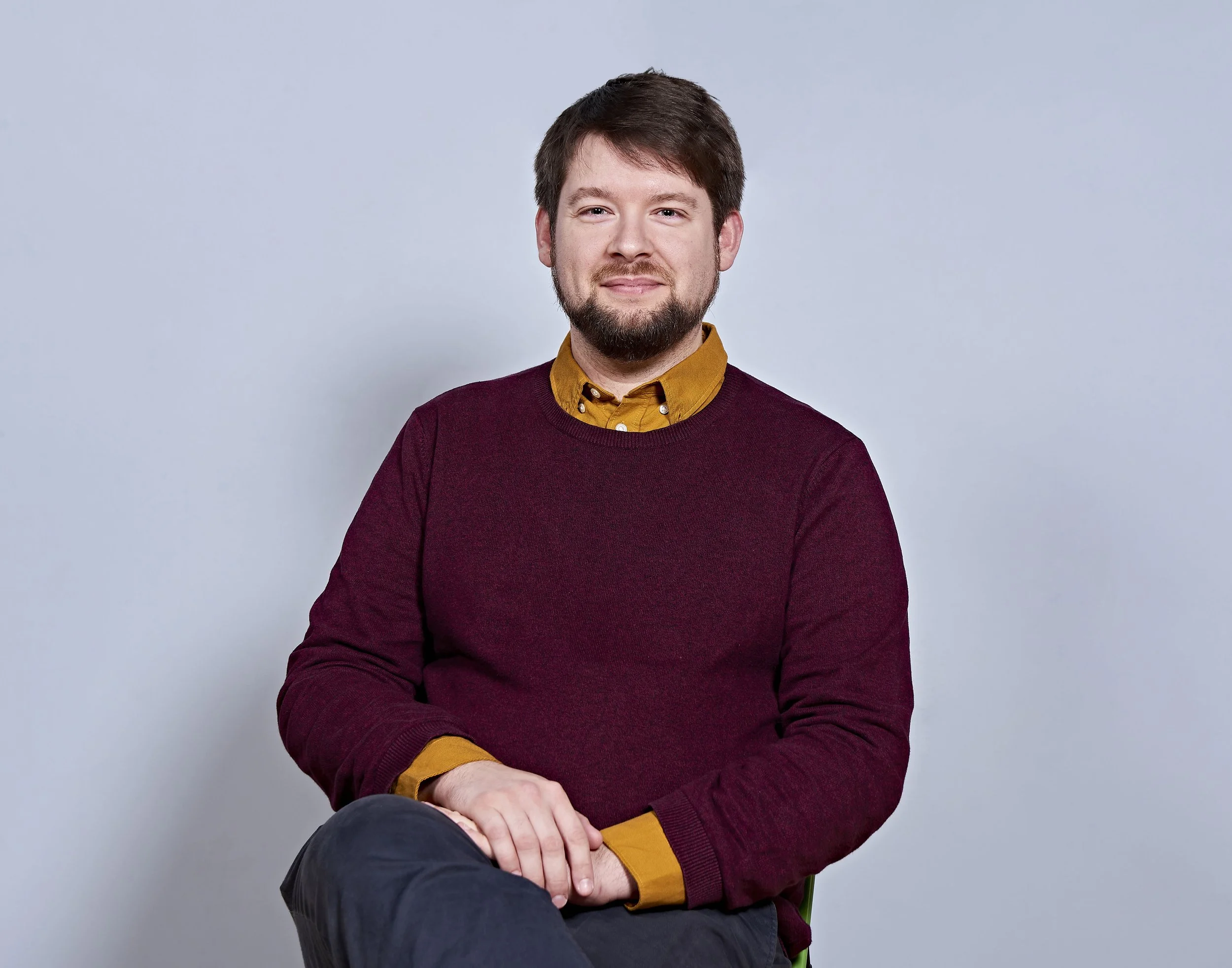 A man sitting and facing the camera, wearing a burgundy sweater and a yellow shirt, against a plain background.