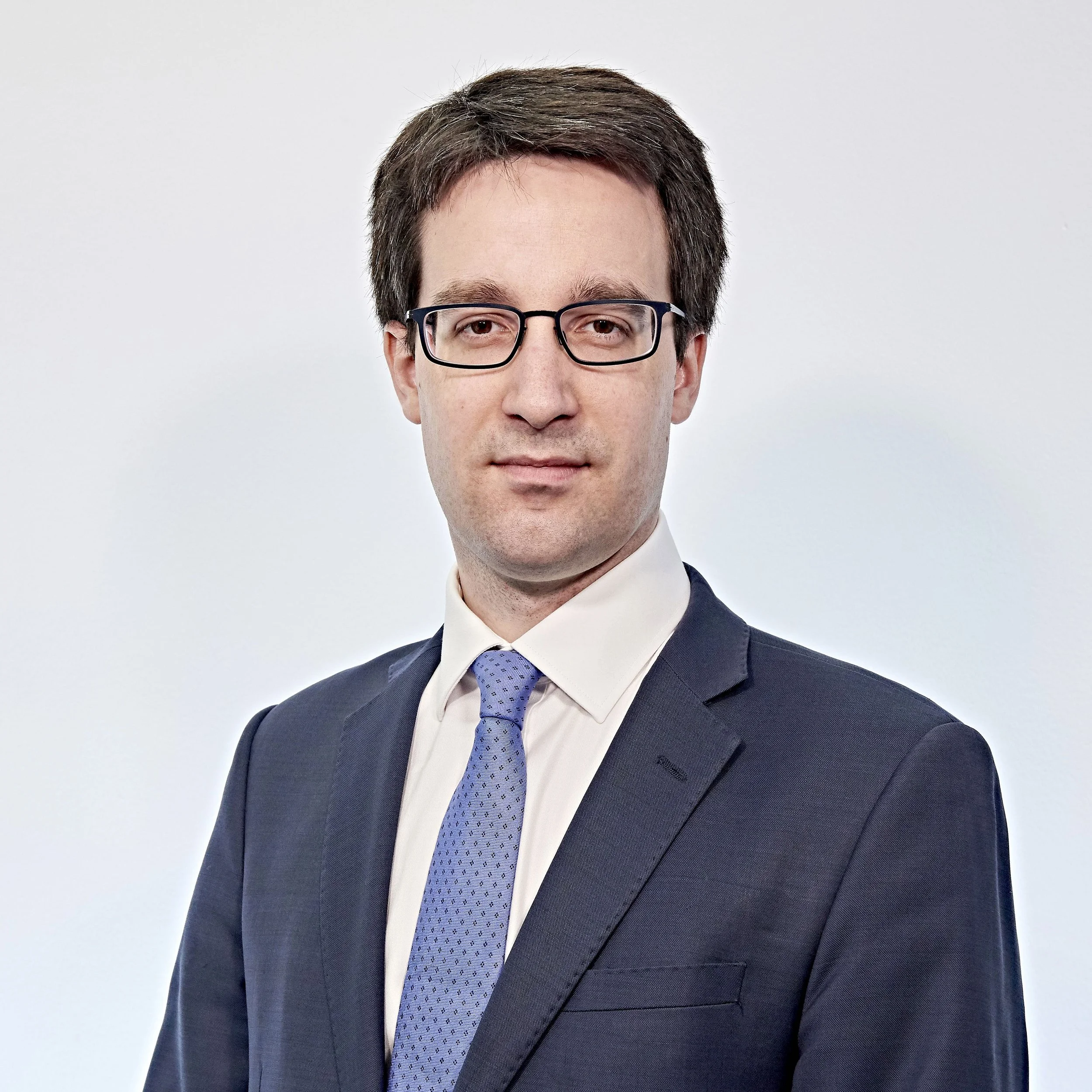 Professional man wearing glasses and a suit with a blue tie against a plain background.