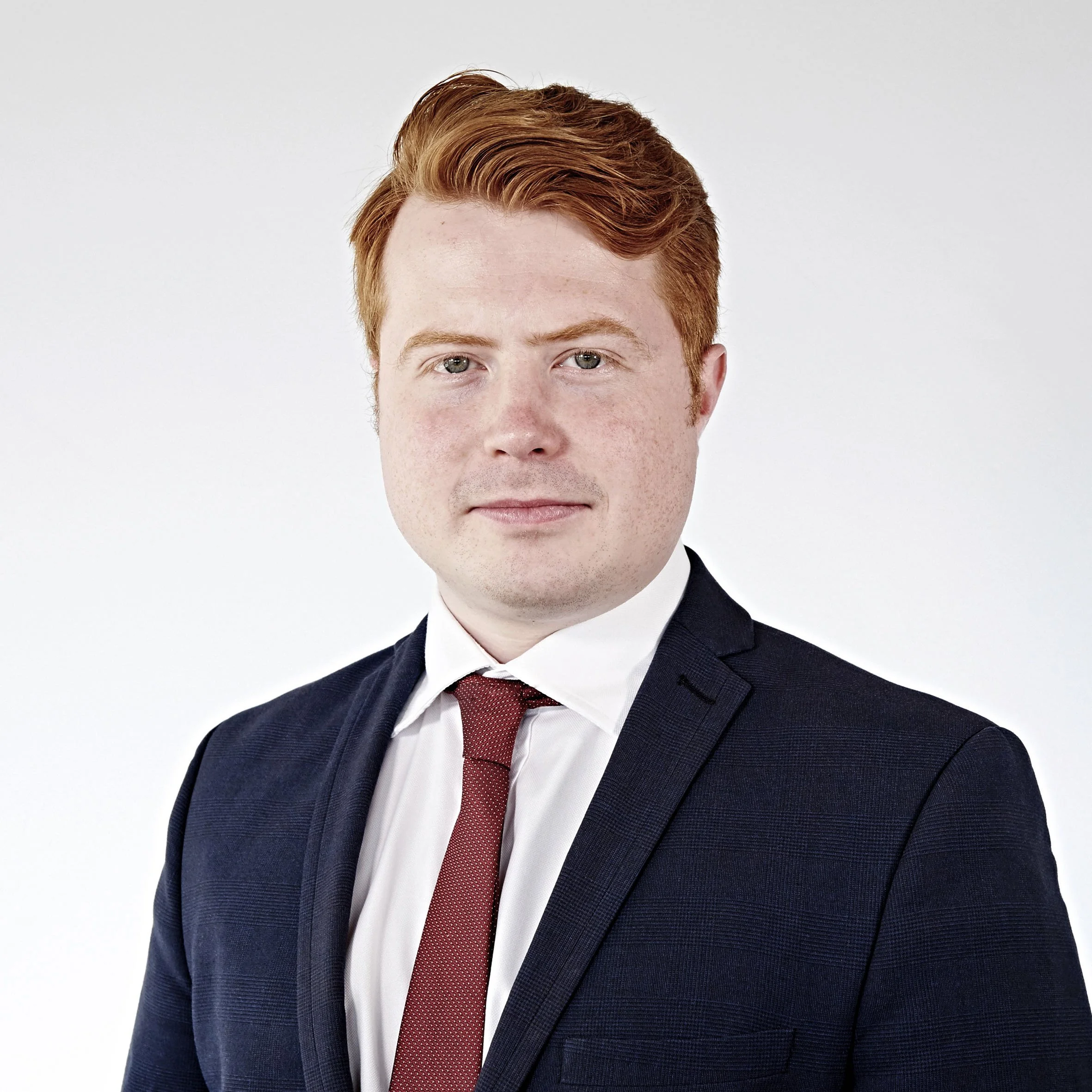 A man with red hair wearing a navy suit and red tie is posing against a plain white background.