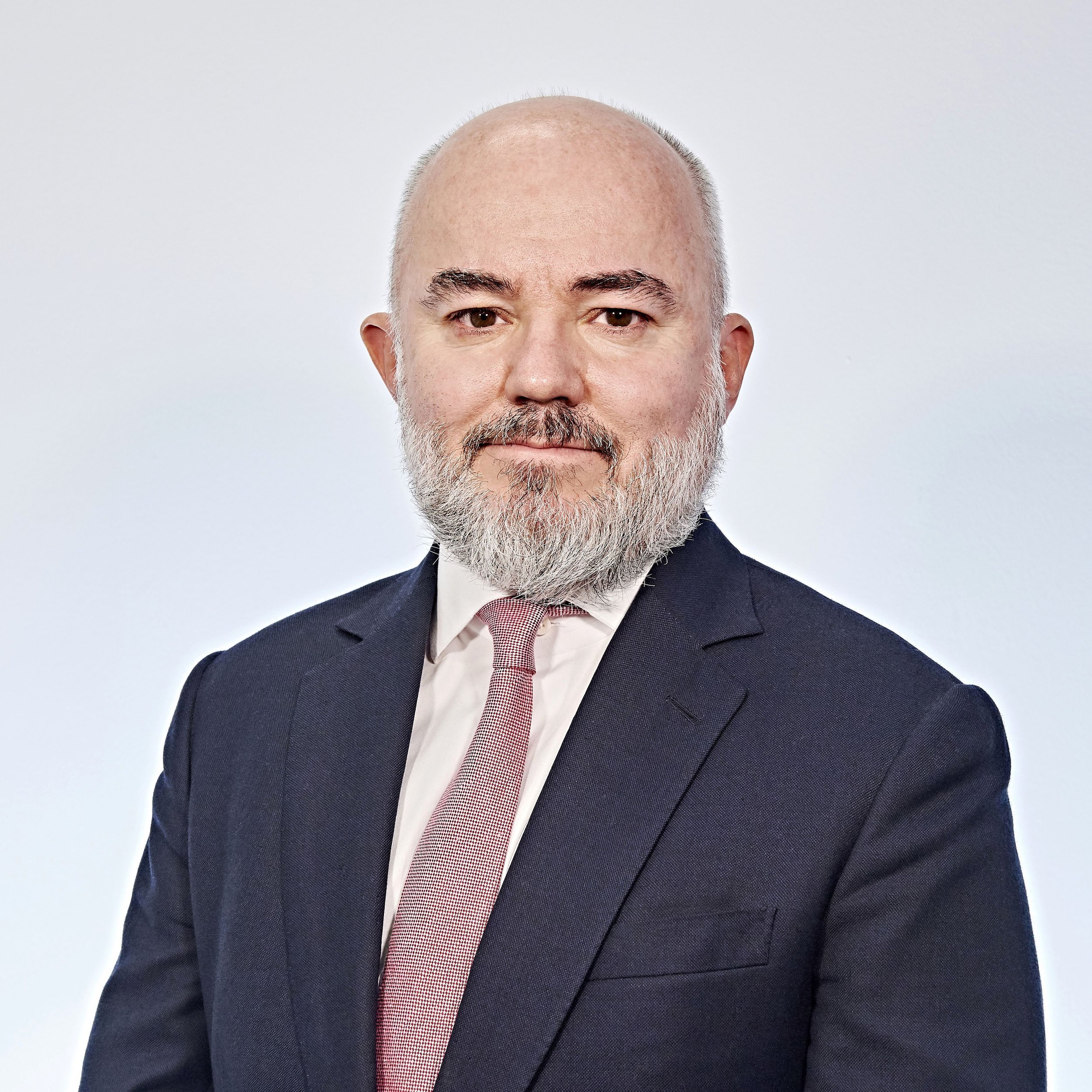 A bald man with a beard wearing a suit and tie, posing against a plain background.
