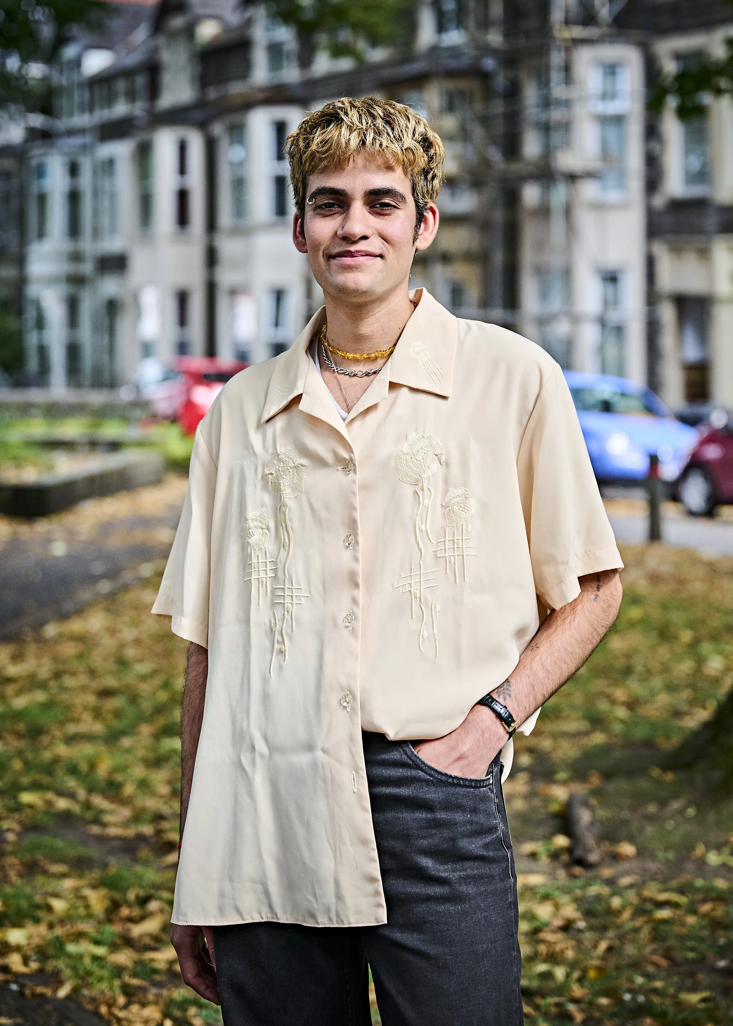 A person wearing a light-colored short-sleeve shirt embroidered with floral patterns stands outdoors in front of a row of buildings. The person has short, light brown hair and wears a necklace and a watch. The background includes parked cars and tree