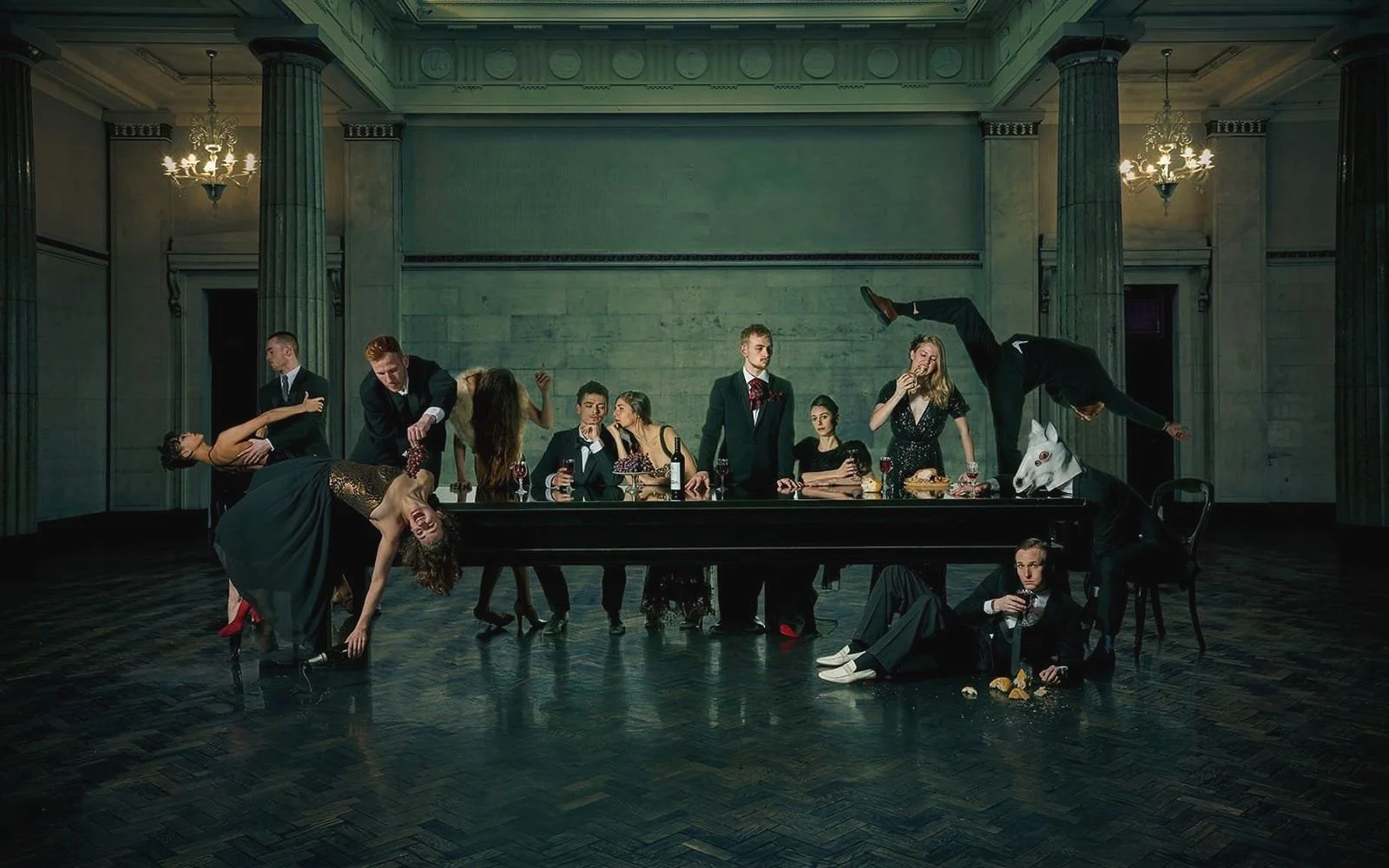 A large group of people dressed in formal attire sit and stand around a long dining table in a grand room with marble walls and tall columns. Some individuals are engaged in lively or dramatic poses, including a man with a white horse mask and a woman with a wine bottle. The scene appears theatrical or surreal.