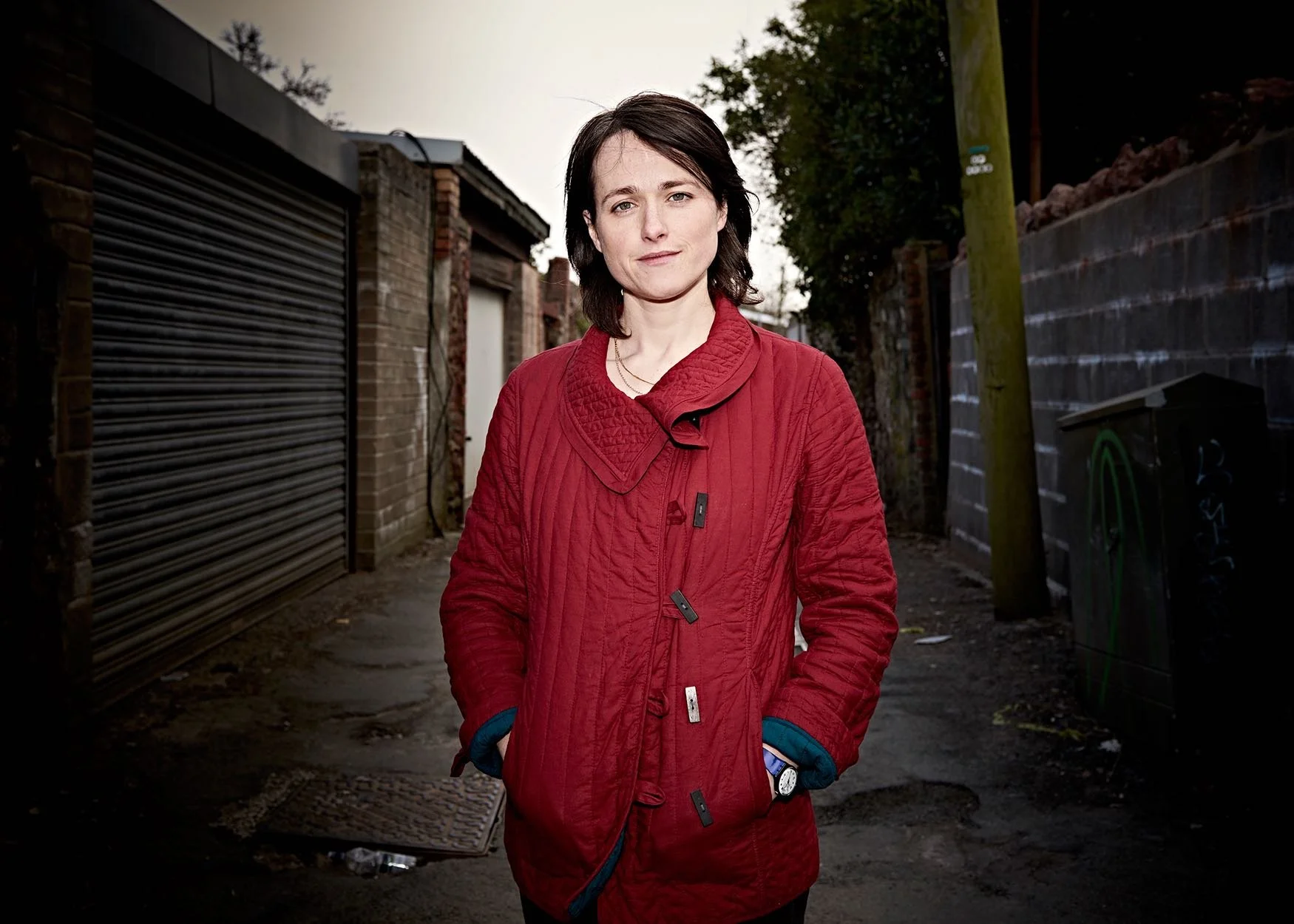 A woman with shoulder-length dark hair wearing a red quilted jacket standing outdoors in an alley with brick and wood structures, a wood utility pole, and a trash bin.