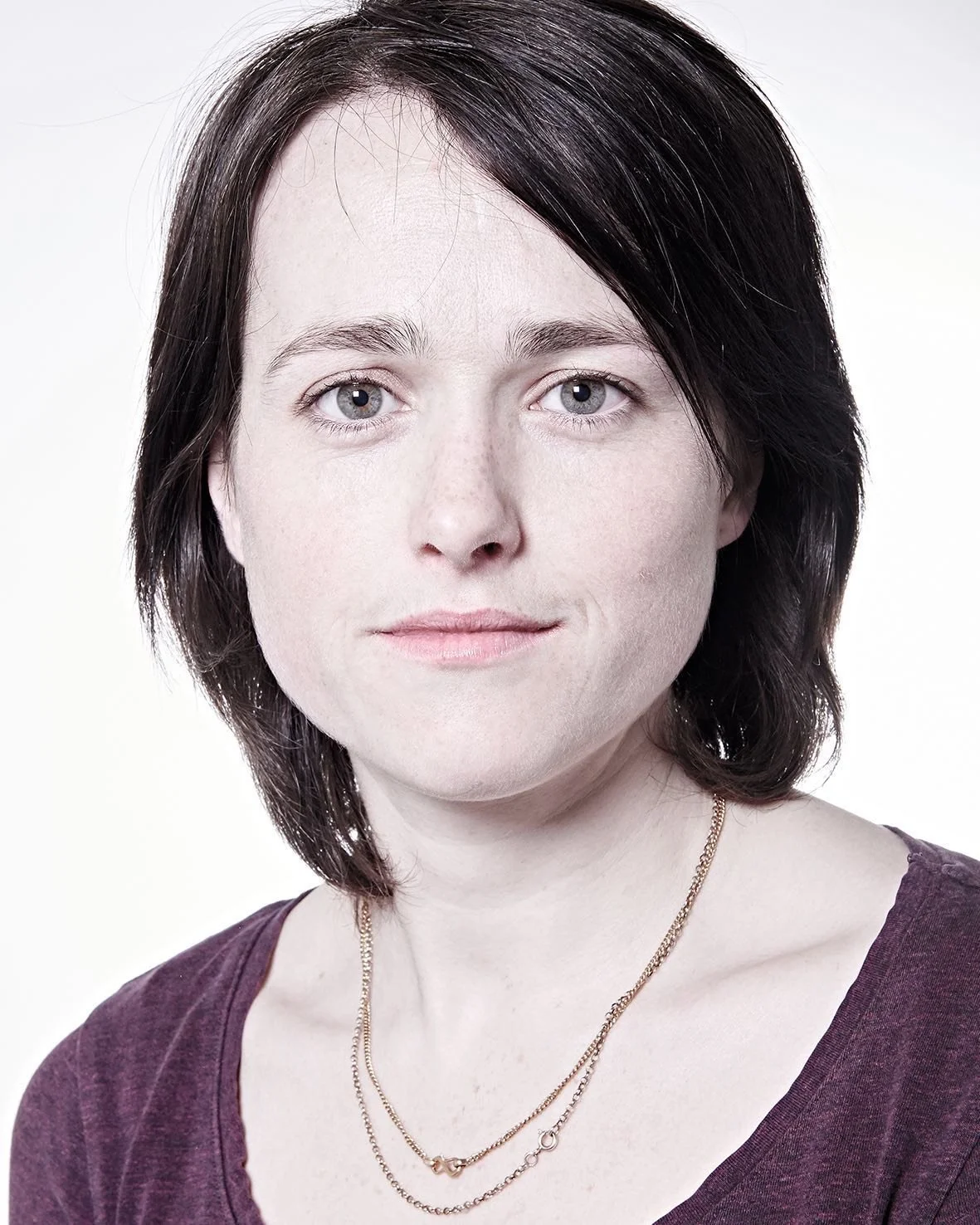 Portrait of a person with short dark hair and a neutral expression, wearing a purple shirt and a gold necklace, against a white background.