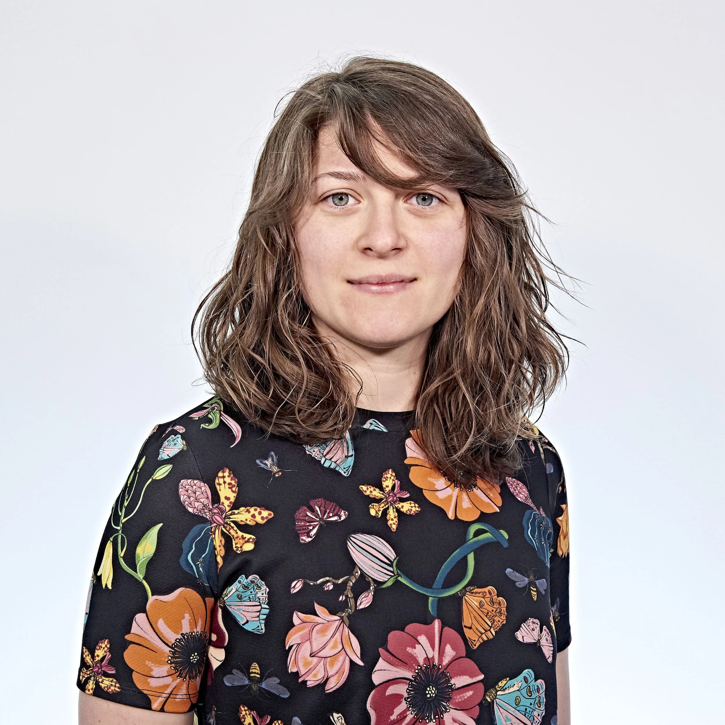 Person with wavy brown hair wearing a floral-patterned top against a plain background.