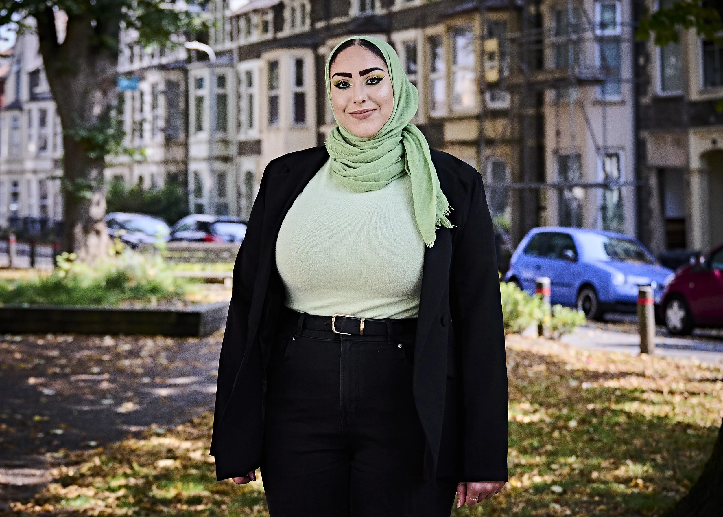Person wearing a green hijab and black jacket standing outdoors with trees and parked cars in the background.