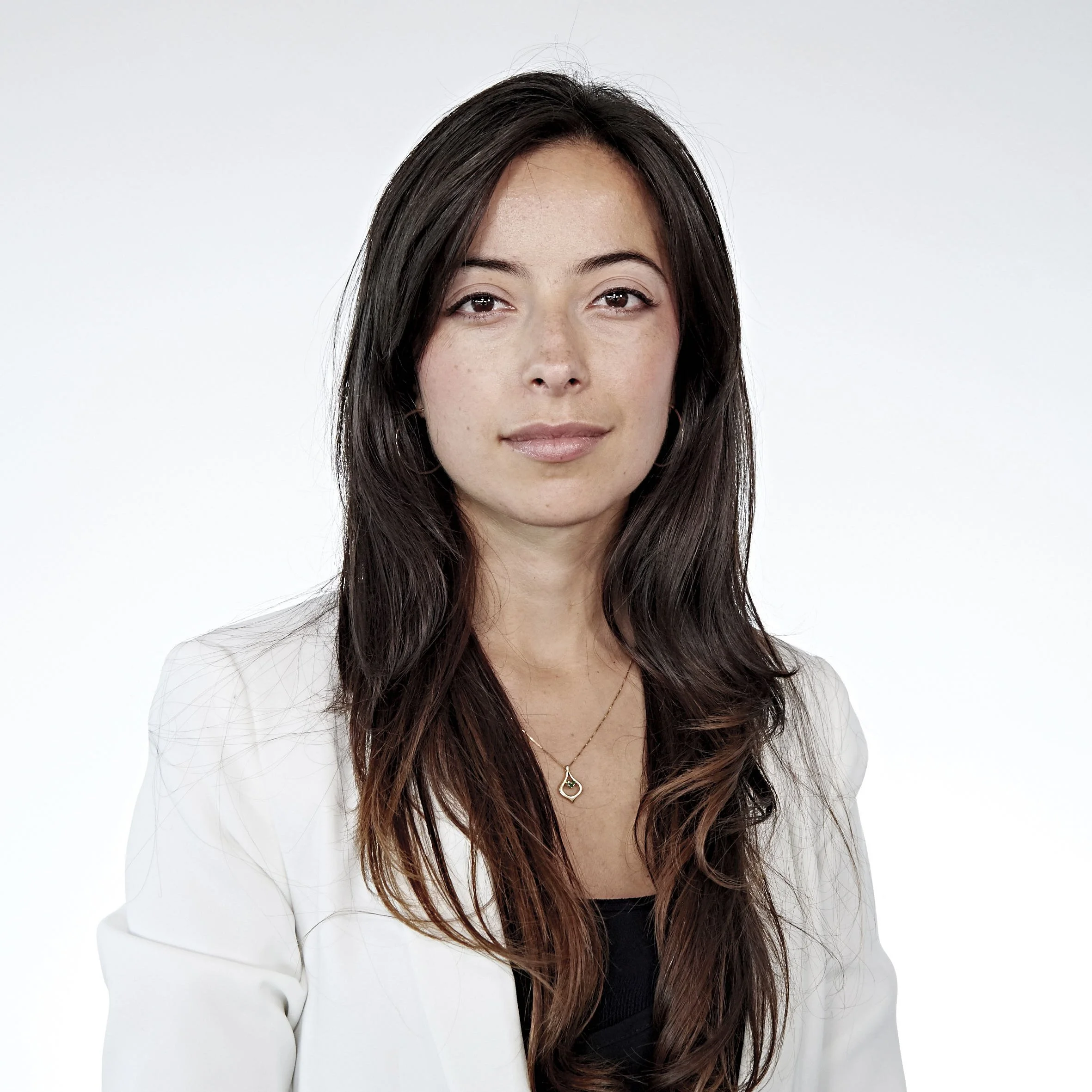 Woman wearing a white blazer and a pendant necklace, with long brown hair, posing against a plain background.