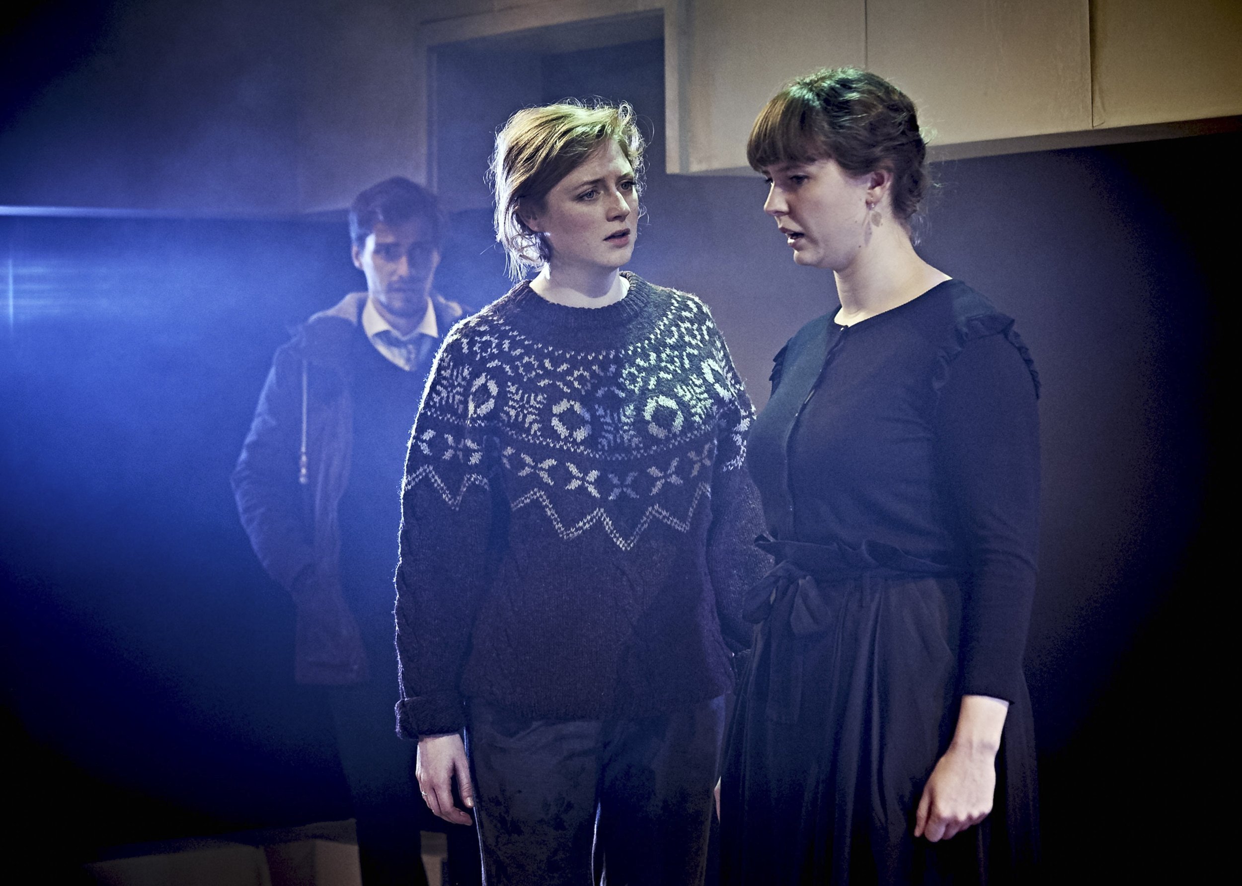 Two women are having an intense conversation, facing each other, with a man in the background. The women are dressed casually, and the scene appears to be indoors with soft lighting.