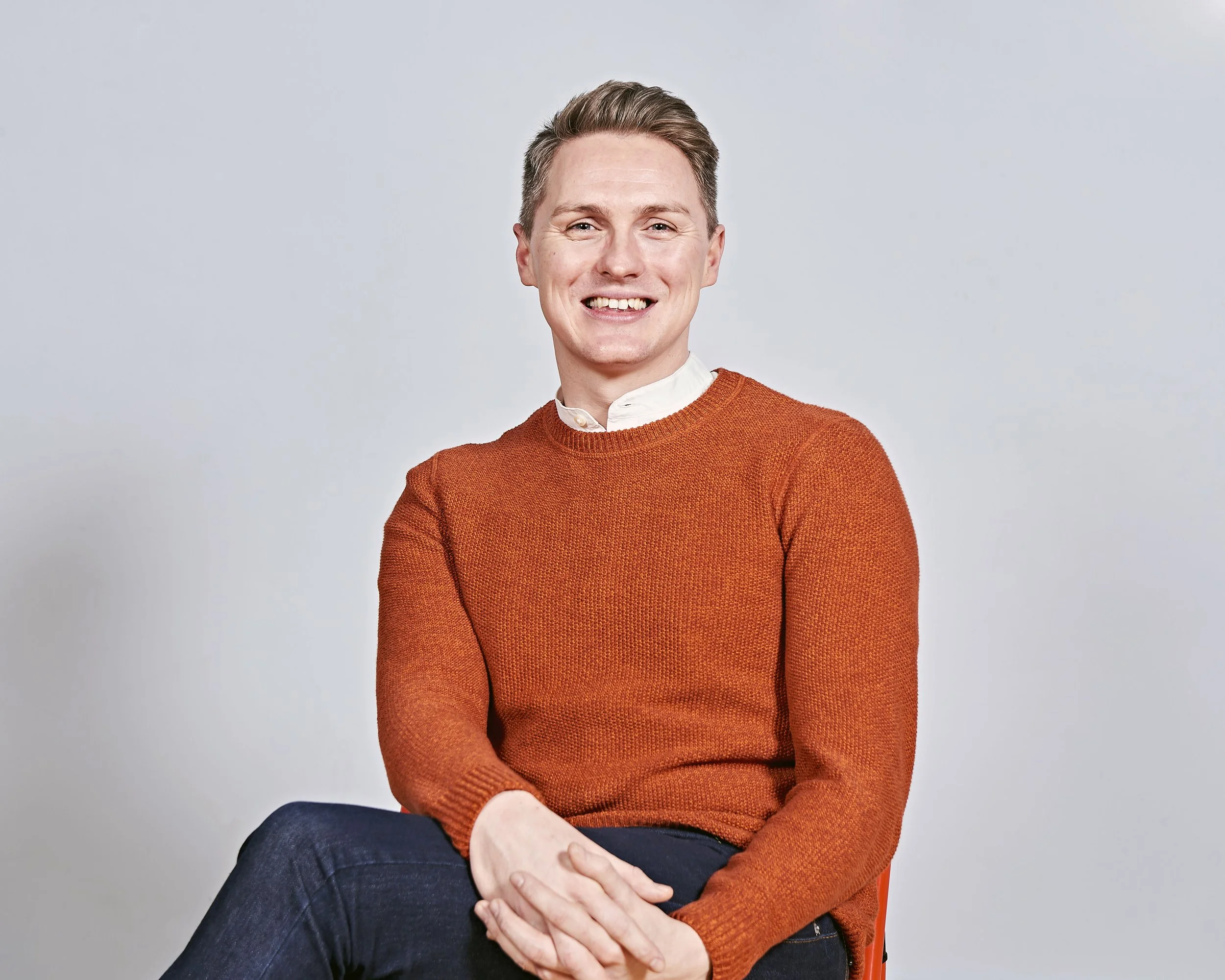 Man wearing an orange sweater sitting on a chair against a plain background, smiling.
