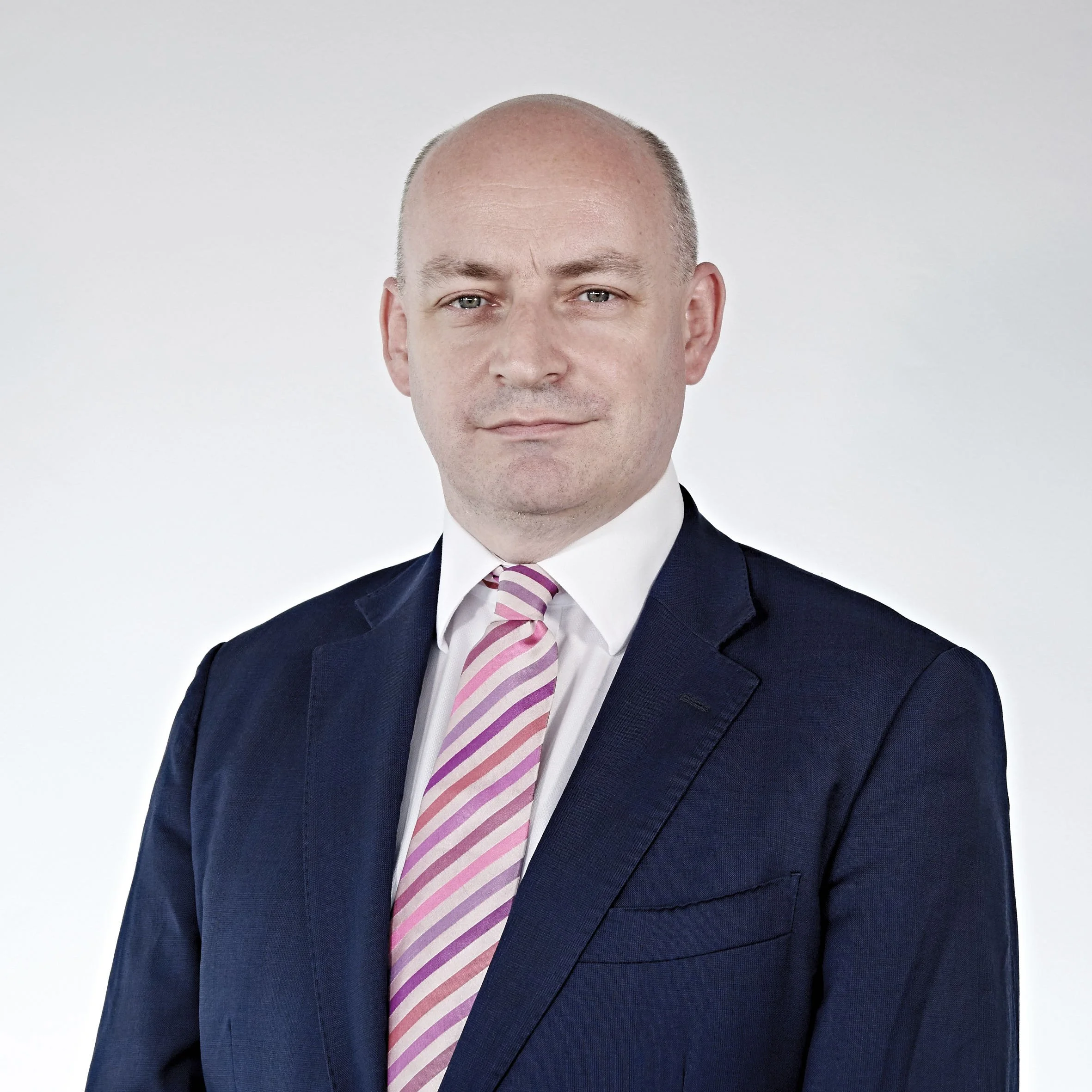 Bald man in a suit with a pink striped tie, posing against a plain background.