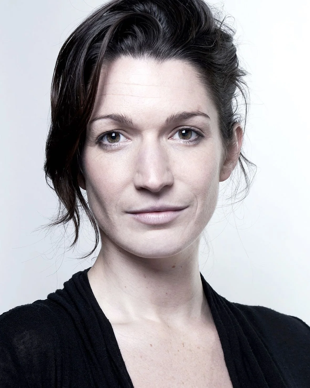 Close-up portrait of a woman with short dark hair, wearing a black top, looking directly at the camera against a light background.