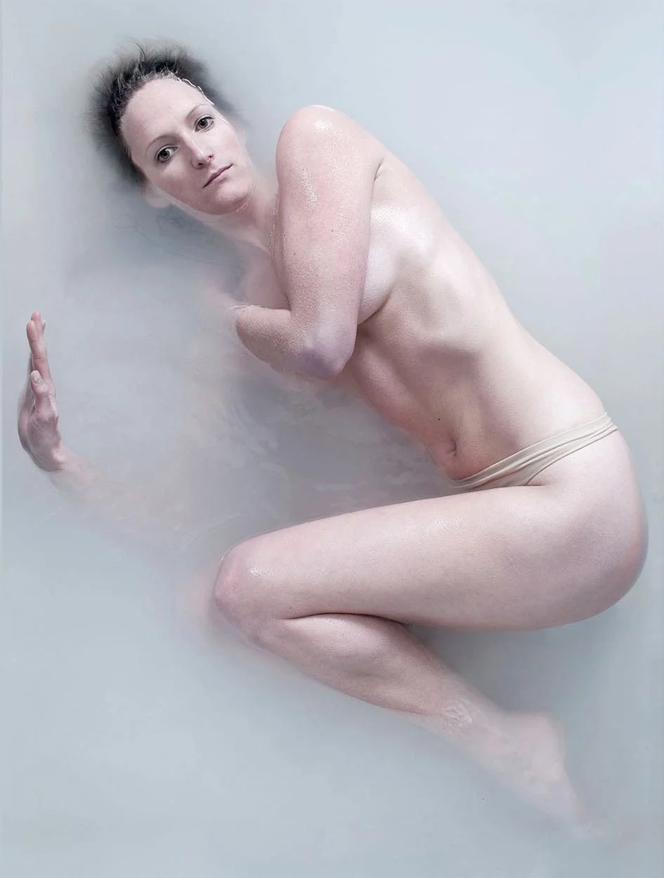A woman with dark hair submerged in milky water, with her head tilted back and arms positioned protectively. She is wearing no clothes and is covered in a thin layer of water or sweat.