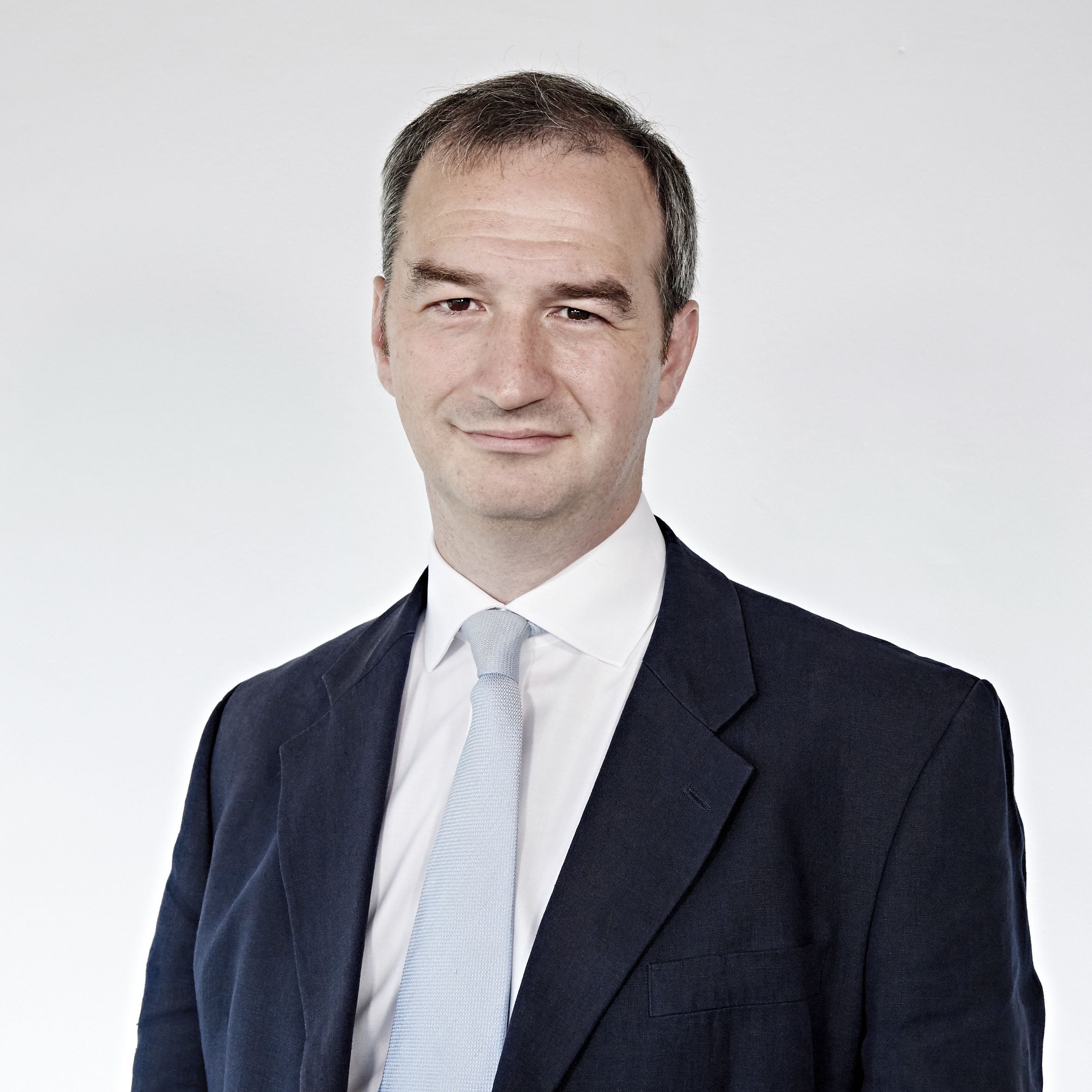 A man in a dark suit and light tie against a neutral background.