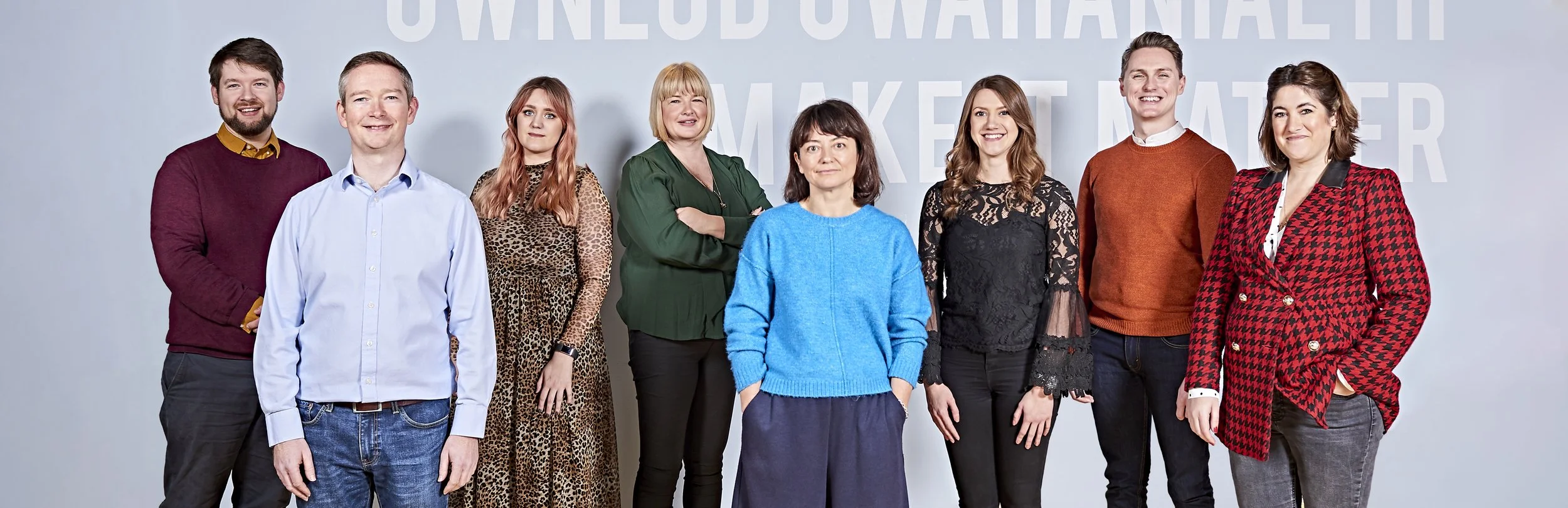 A group of nine people standing against a light-colored background featuring text. They are dressed in various styles, with some wearing casual attire and others in more formal outfits. The group includes both men and women, each positioned to face t