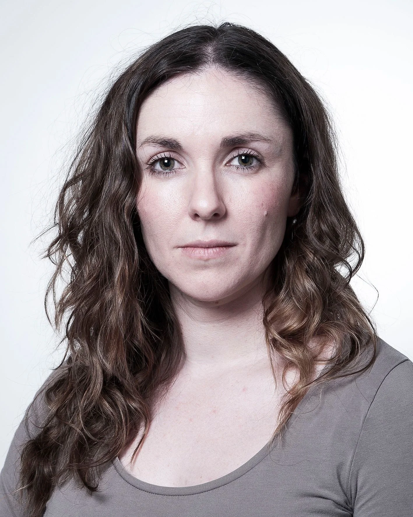 Woman with brown wavy hair and a neutral expression, wearing a gray top, facing forward.