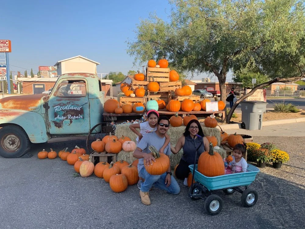 Family at pumpkin patch with pumpkins on truck and hay bales, children in wagon, autumn scene.
