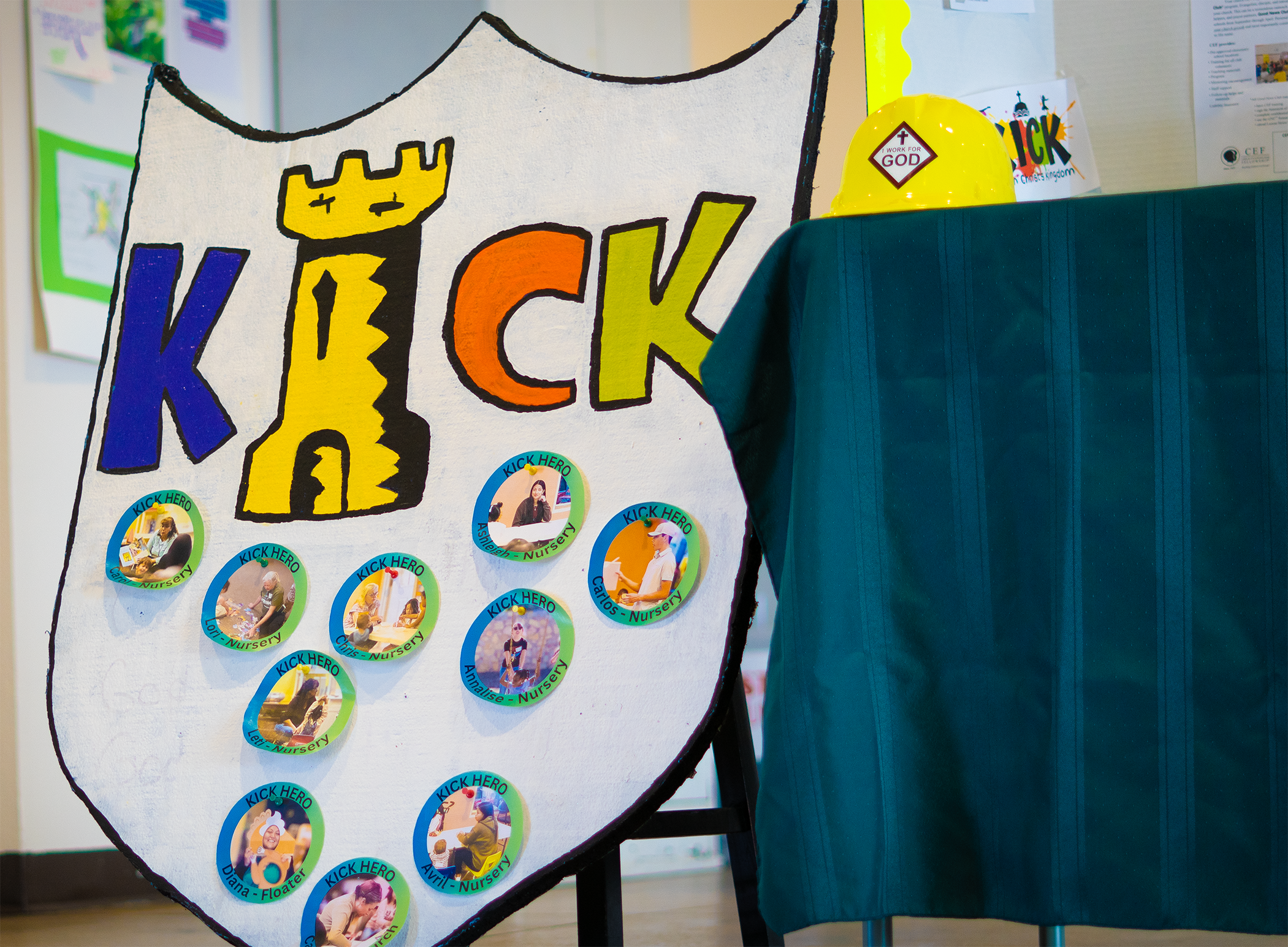 A colorful bulletin board with the word 'KICK' at the top, decorated with photos of children and staff members, each labeled as 'KICK HERO' and by their roles like 'Nursery' or 'Floater', and a yellow safety helmet with a sticker on a table next to the board.