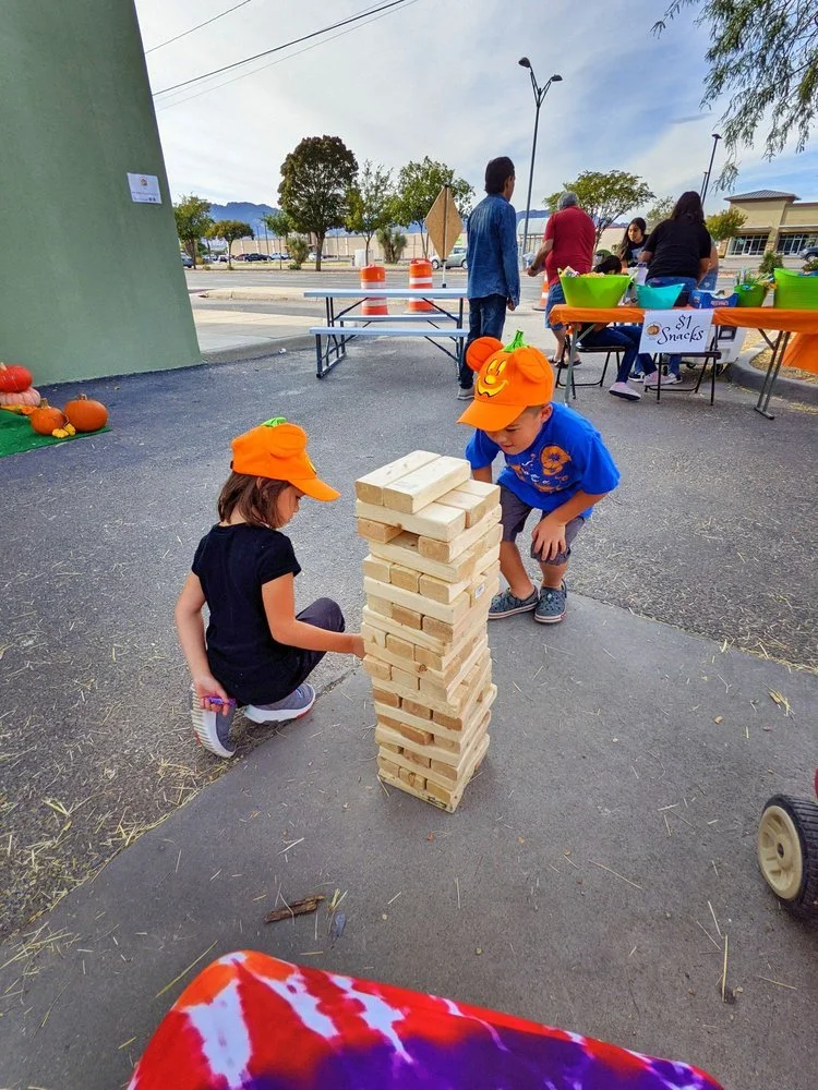 Two children wearing orange pumpkin hats playing giant Jenga outdoors at a fall festival, with pumpkins and a snack table in the background.