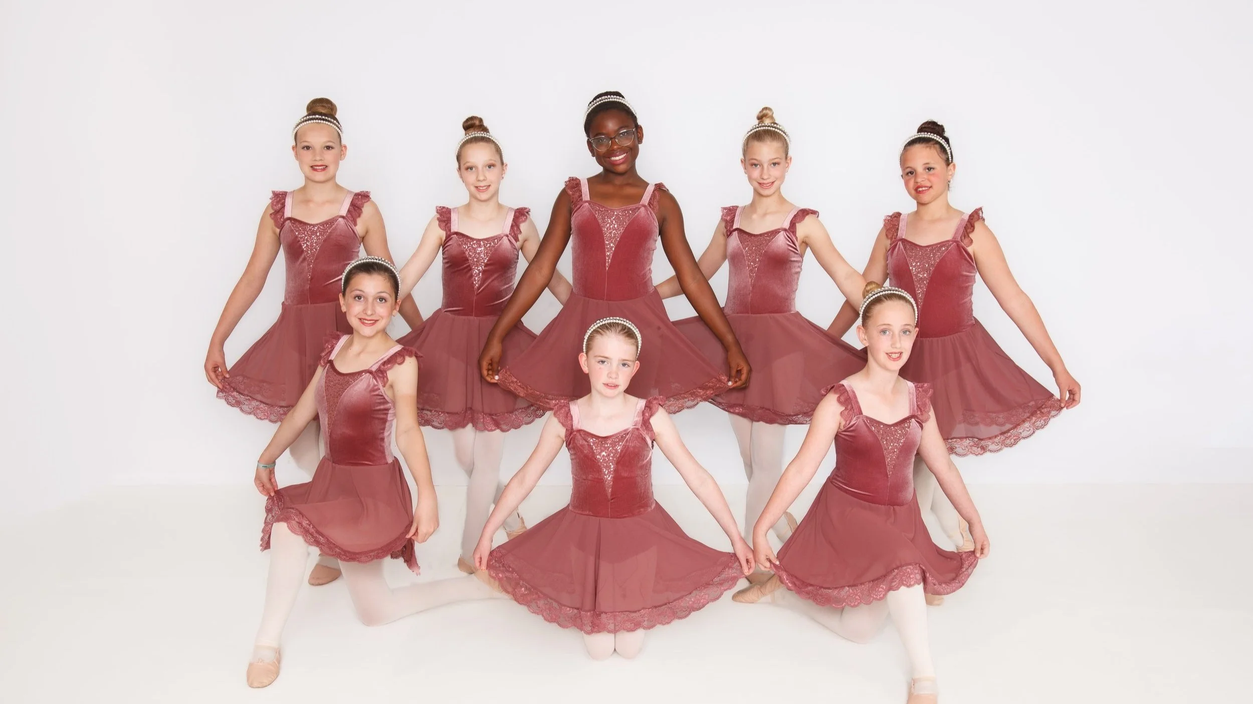 Group of nine young girls in matching ballet costumes and headbands, posing together in a studio with a white background.
