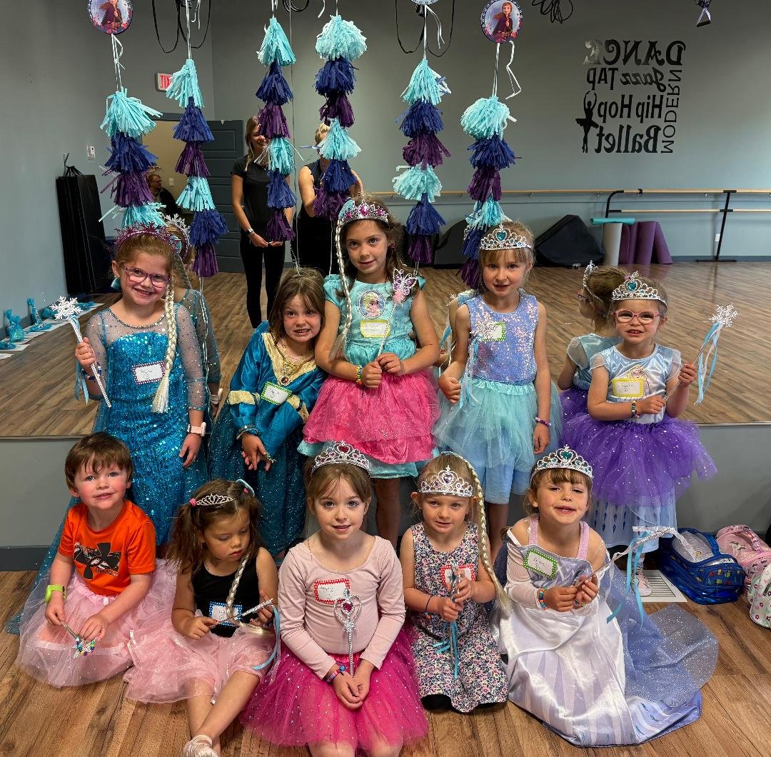Children dressed as princesses and fairies with tiaras, holding wands, in a dance studio decorated with purple, teal, and blue tissue paper pom-poms hanging from the ceiling.