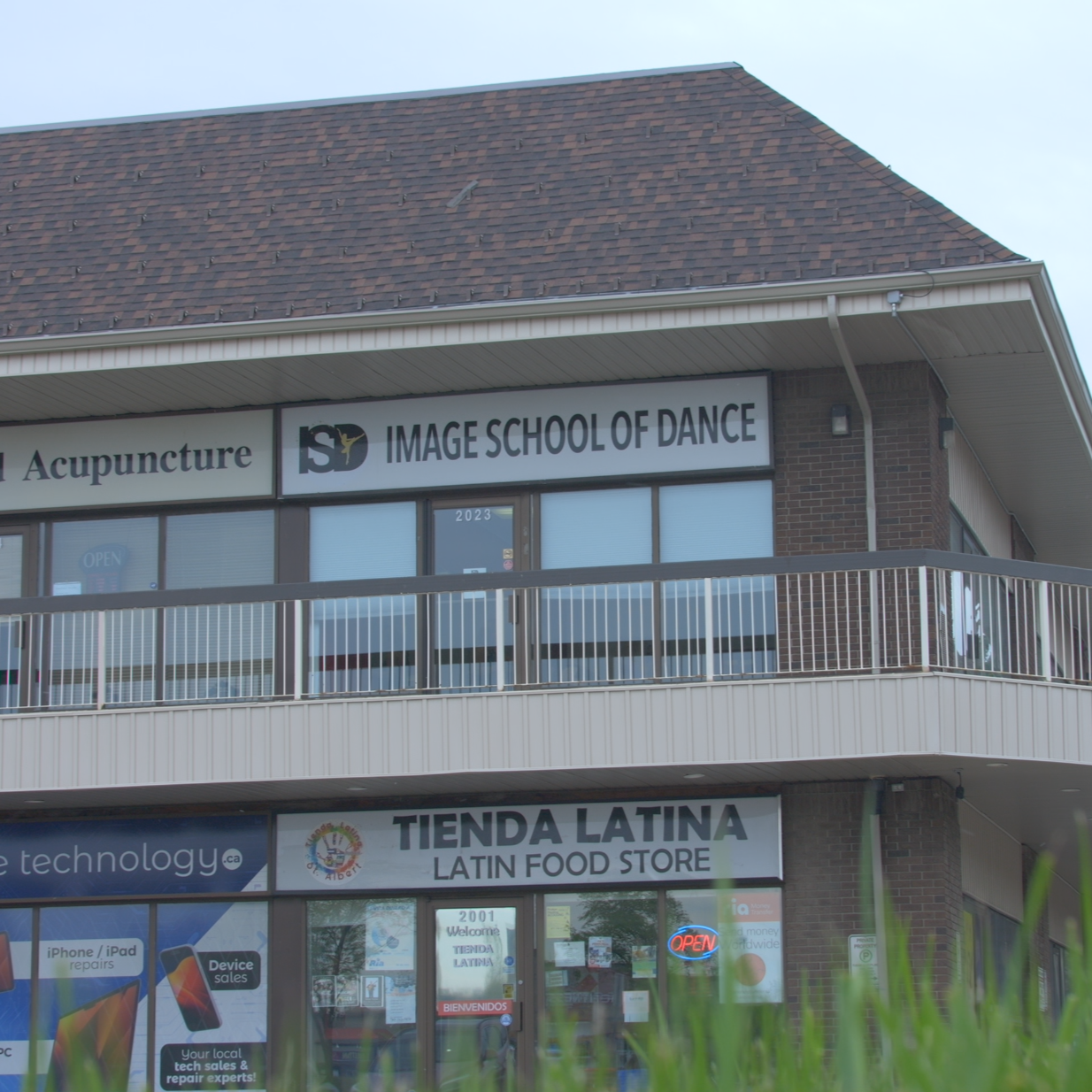 A two-story commercial building with signs for 'IMAGE SCHOOL OF DANCE' and 'TIENDA LATINA LATIN FOOD STORE'. The dance school sign is on the upper level and includes a logo with a person dancing. The food store sign is on the lower level with a colorful logo. There are large windows on both levels, with some signs and posters visible inside. The building has a tan facade with brick accents, and the roof is covered with brown shingles.