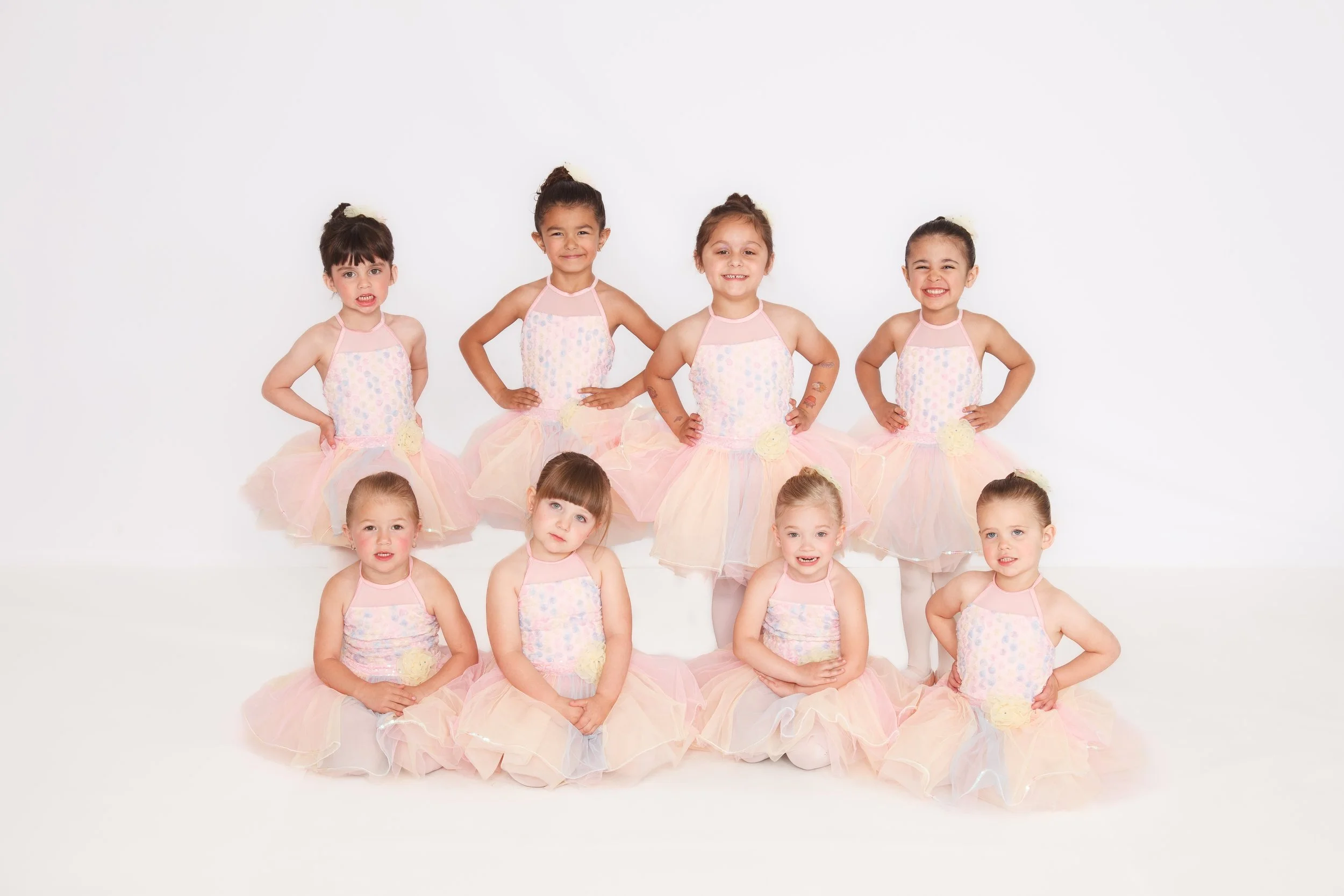 Group of nine young girls in pastel ballet costumes posing against a white background, some standing and some sitting on the floor.