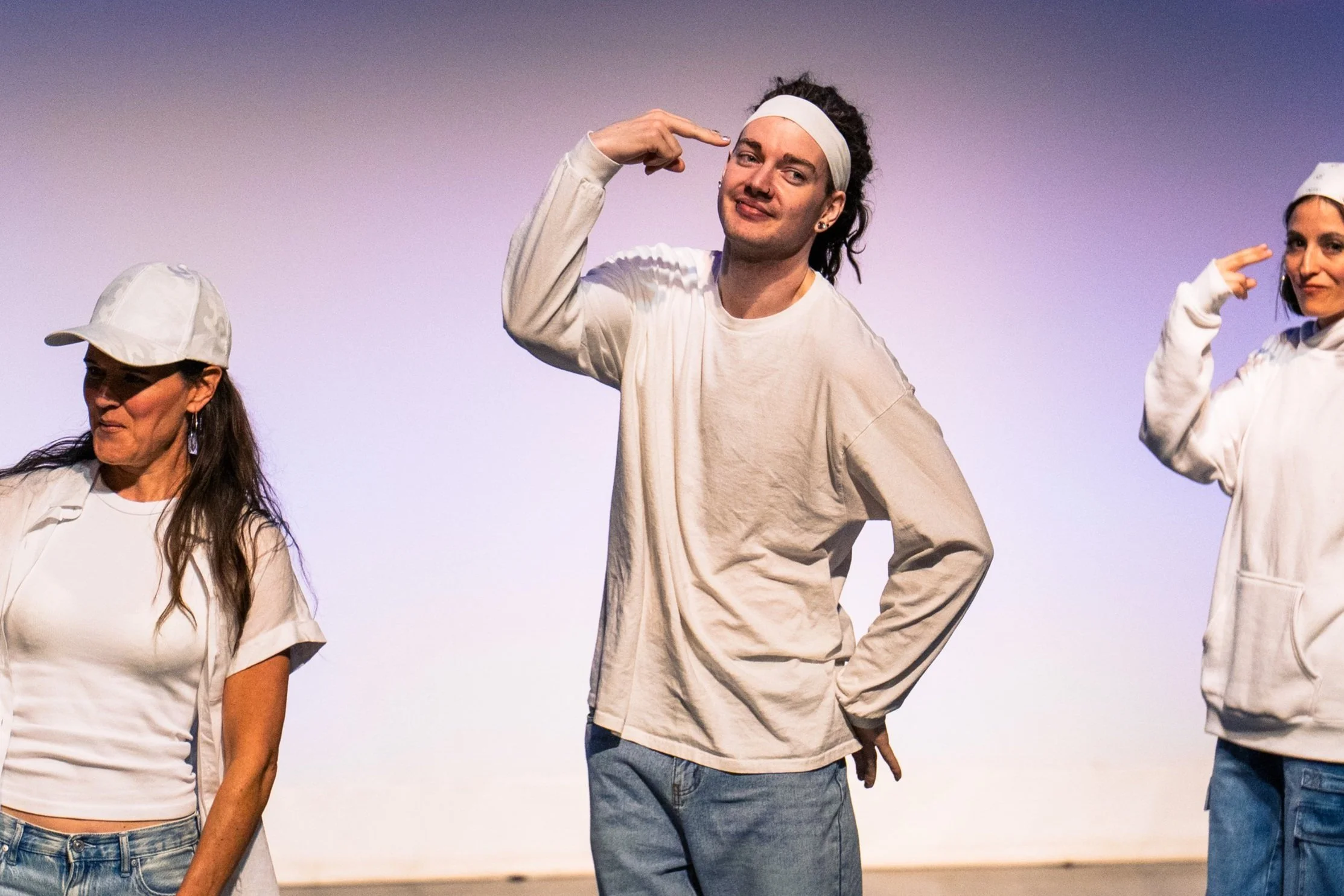 Three people on stage, two women and one man, dressed casually in white and light-colored clothing. They are striking playful poses, with the man in the center touching his head with one hand and the woman on the right making a gesture near her head.